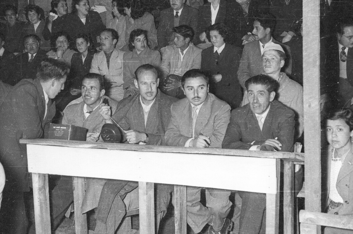 Plano entero de un grupo de hombres sentados. jueces del partido. Uno de ellos está mirando hacia la cámara mientras que otro conversa con un Hombres de pie y el resto mira hacia la cancha. detrás de ellos hay un grupo de Hombres y Mujer sentados en las graderías mientras se llevan a cabo partidos de baloncesto femenino de noche.