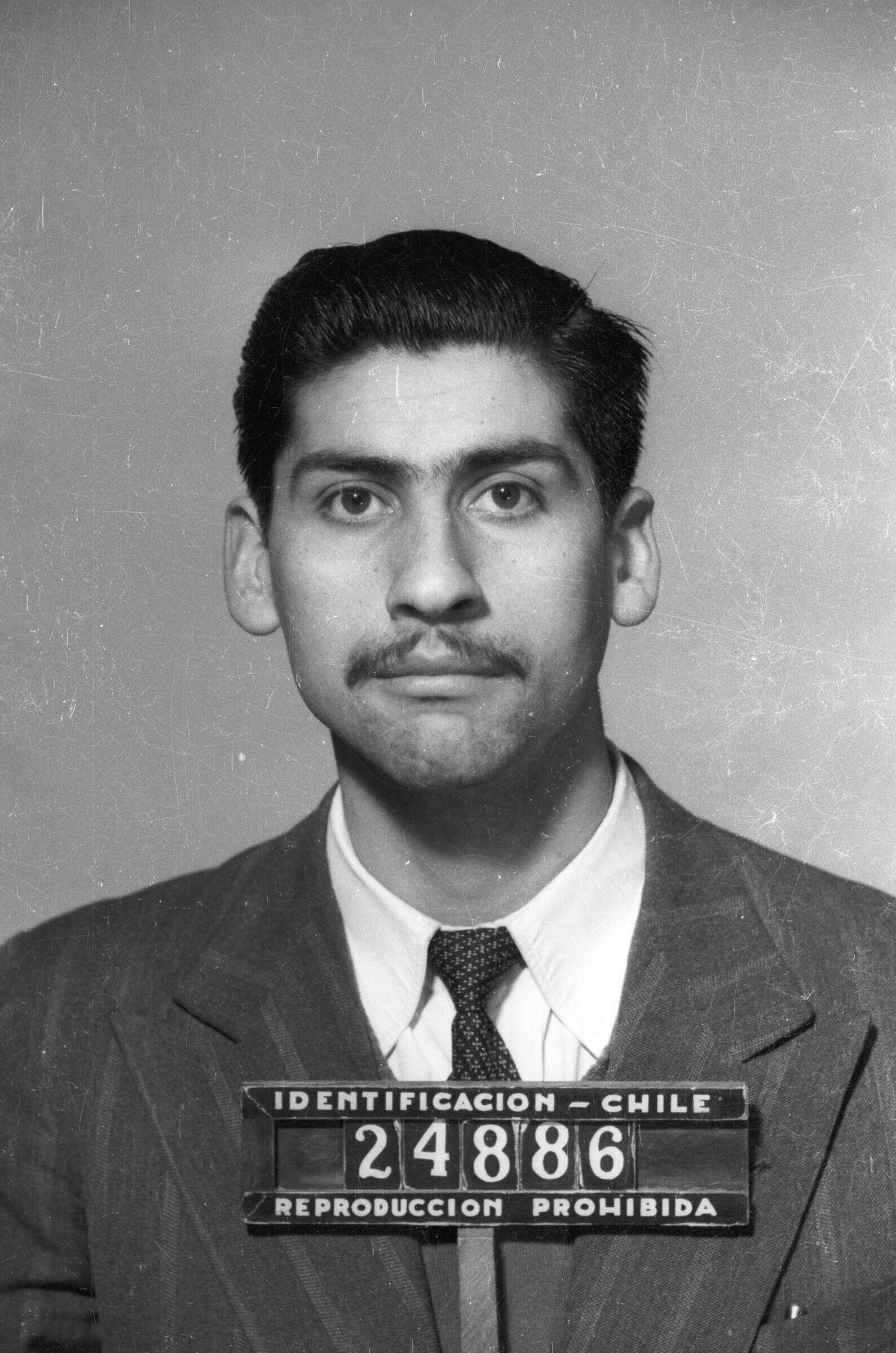 Retrato de estudio de Plano medio de un hombre de Traje posando de frente, sosteniendo un letrero con la inscripción -identificación - Chile 24886 Reproducción Prohibida-