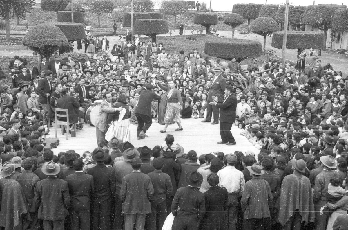 vista superior de plano general de escenario con músicos y dos parejas bailando puesto en una plaza, con gran cantidad de gente rodeando el escenario, lugar no identificado