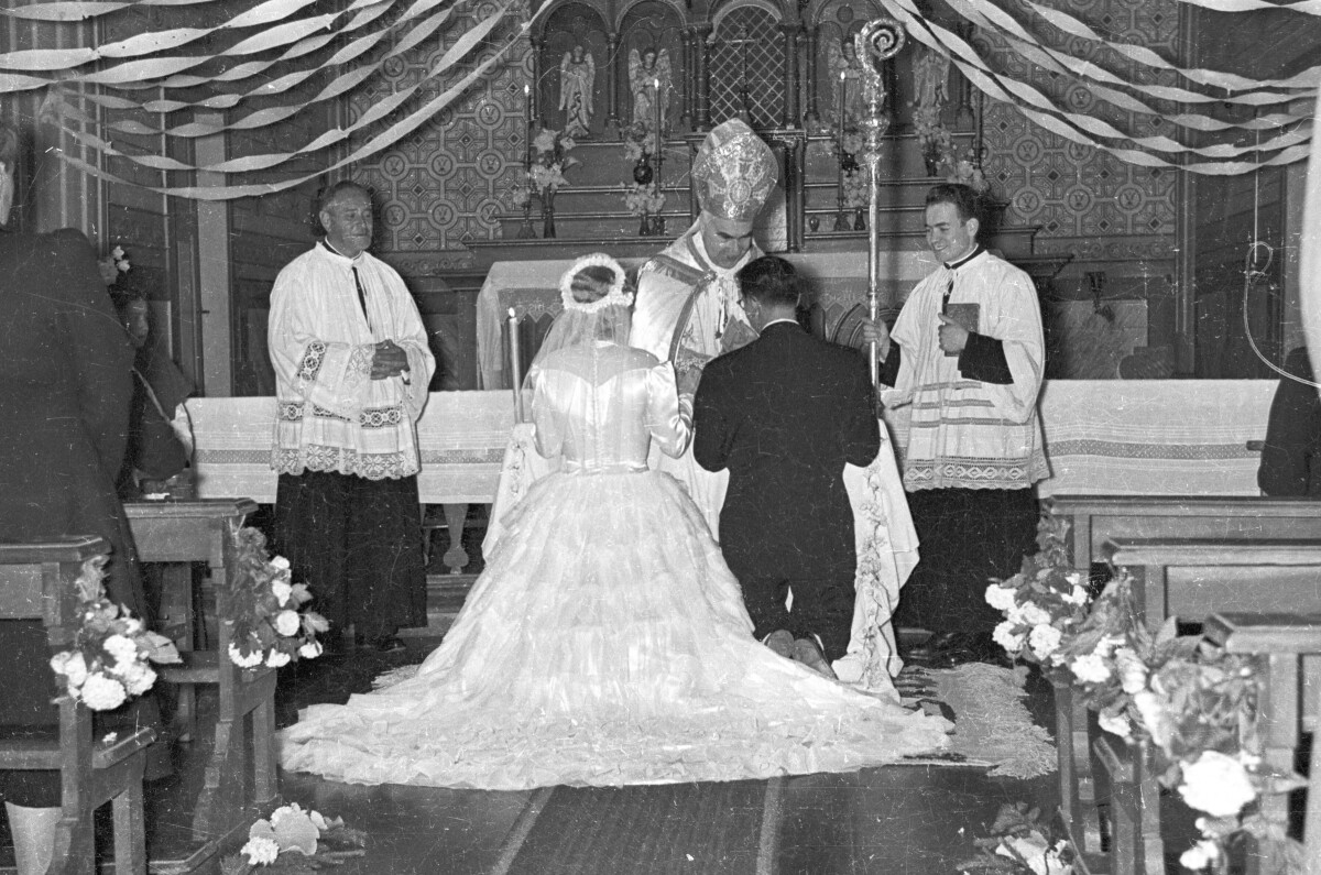 plano general de una pareja de novios, arrodillados en el altar frente a un sacerdote, el cual está acompañado por dos acólitos. Está llevando a cabo su casamiento en el interior de una parroquia no identificada