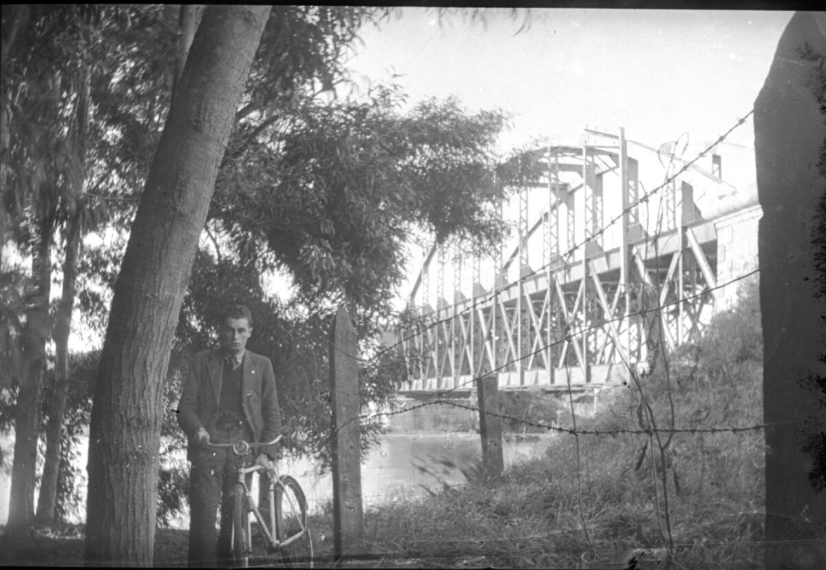 Plano general de un hombre joven posando junto a su bicicleta a orillas de un río atravesado por un puente ferroviario