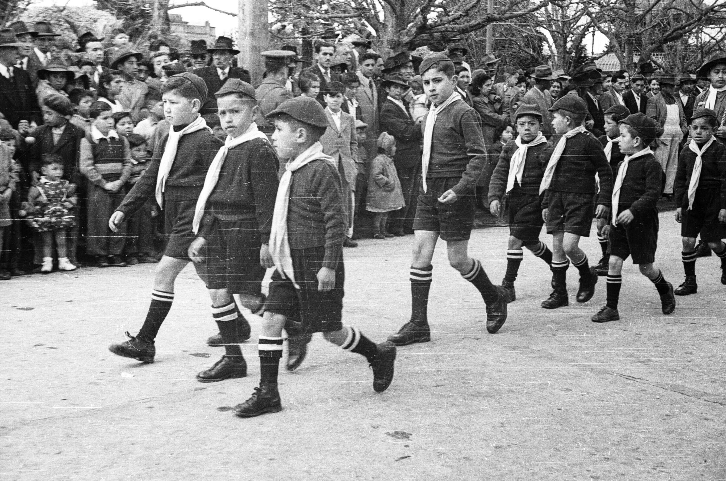 plano general de un grupo de niños caminando en hileras, todos llevan puesto un sombrero, un pañolin y calcetas largas con dos franjas blancas. En un costado de la calle hay un grupo número de Hombres, Mujeres, Niños y Niñas observándolos de pie sobre la vereda durante un desfile por las calles de La Unión