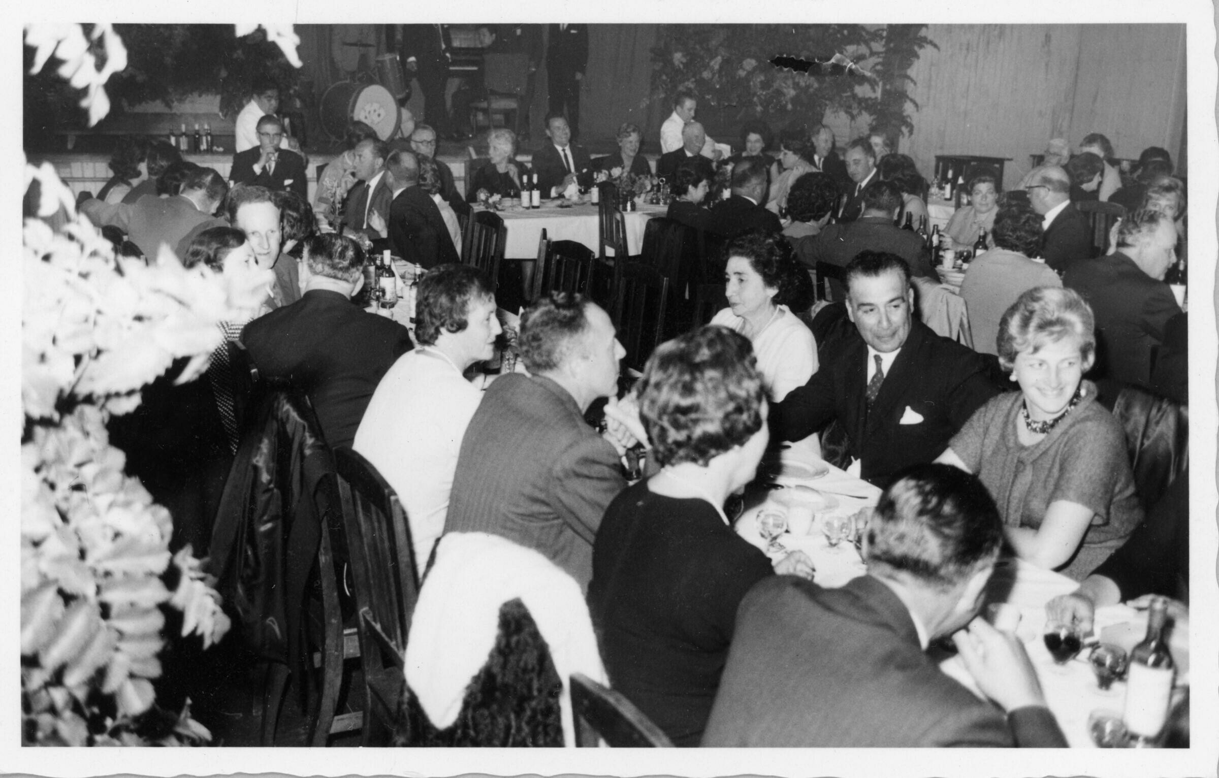 plano general de un grupo de Hombres y Mujeres sentados alrededor de largas mesas mientras comen y conversan entre sí. En el fondo, hay un grupo musical preparándose sobre el escenario y dos garzones que atienden a las personas, parados junto a las mesas. Están en el interior de un local no identificado durante un banquete realizado en honor a Olga Boettcher tras haber finalizado su gobernación