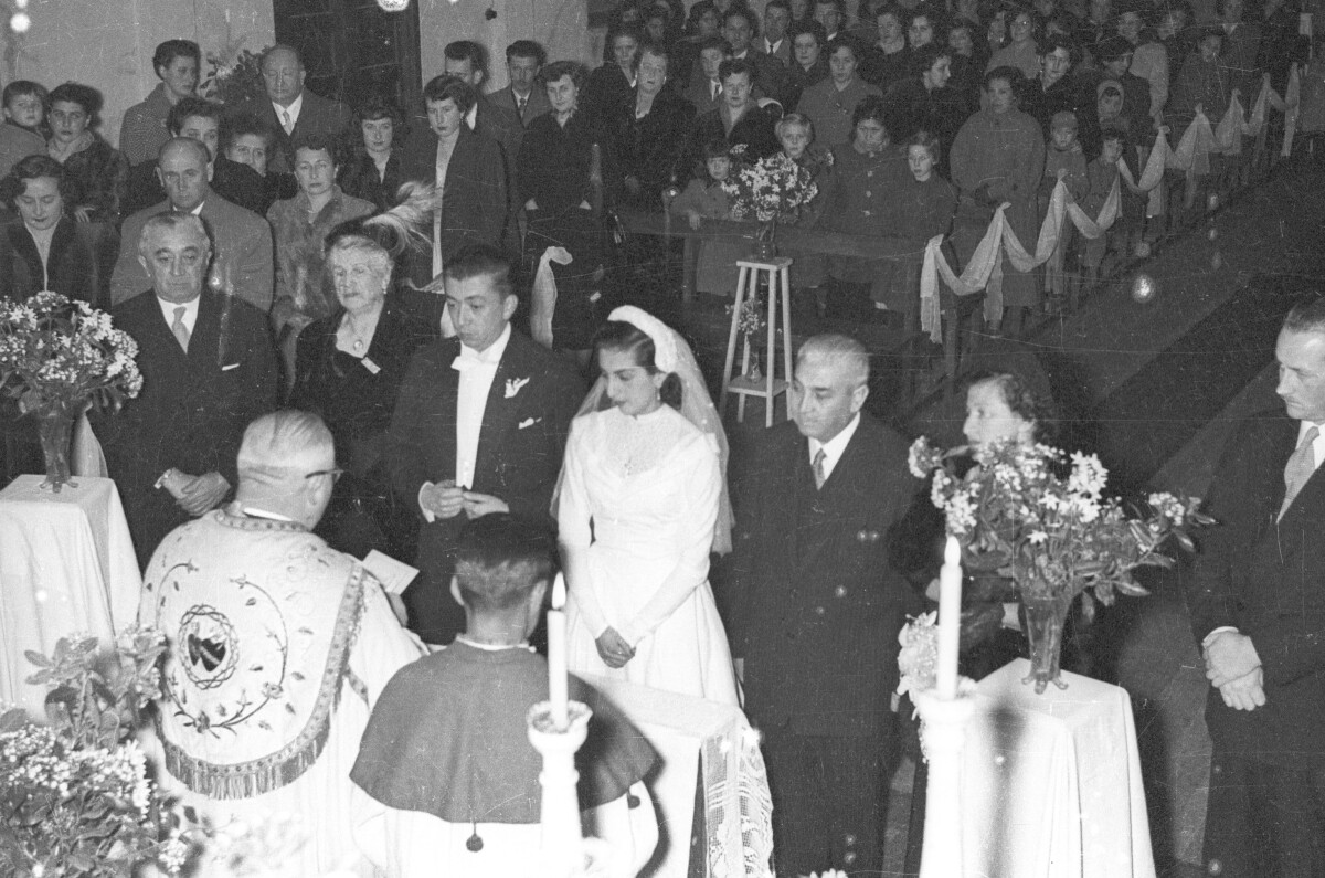 Vista desde un plano alto hacia una pareja de novios y sus padrinos parados frente al altar mientras el sacerdote e dirige hacia ellos. En el fondo están de pie los invitados durante la celebración del sacramento de matrimonio en el interior de una iglesia.