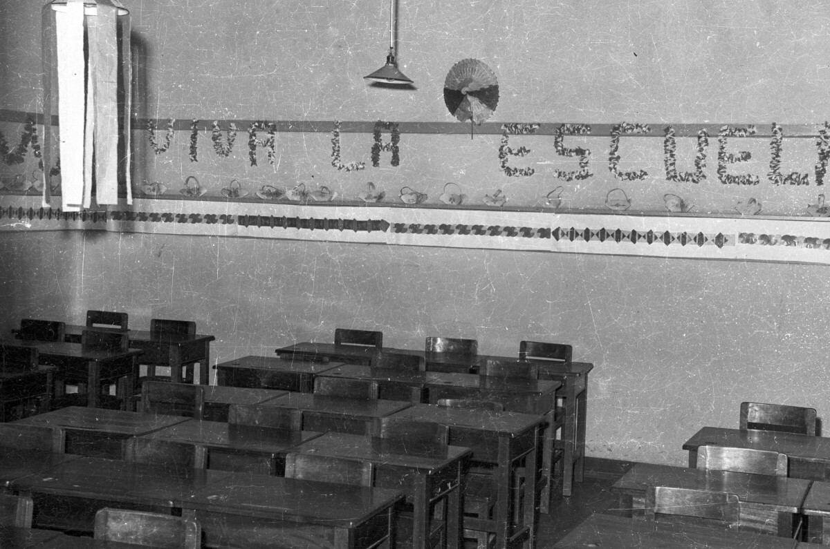 Sala de clases con pupitres y un mensaje de -Viva la escuela- en la pared