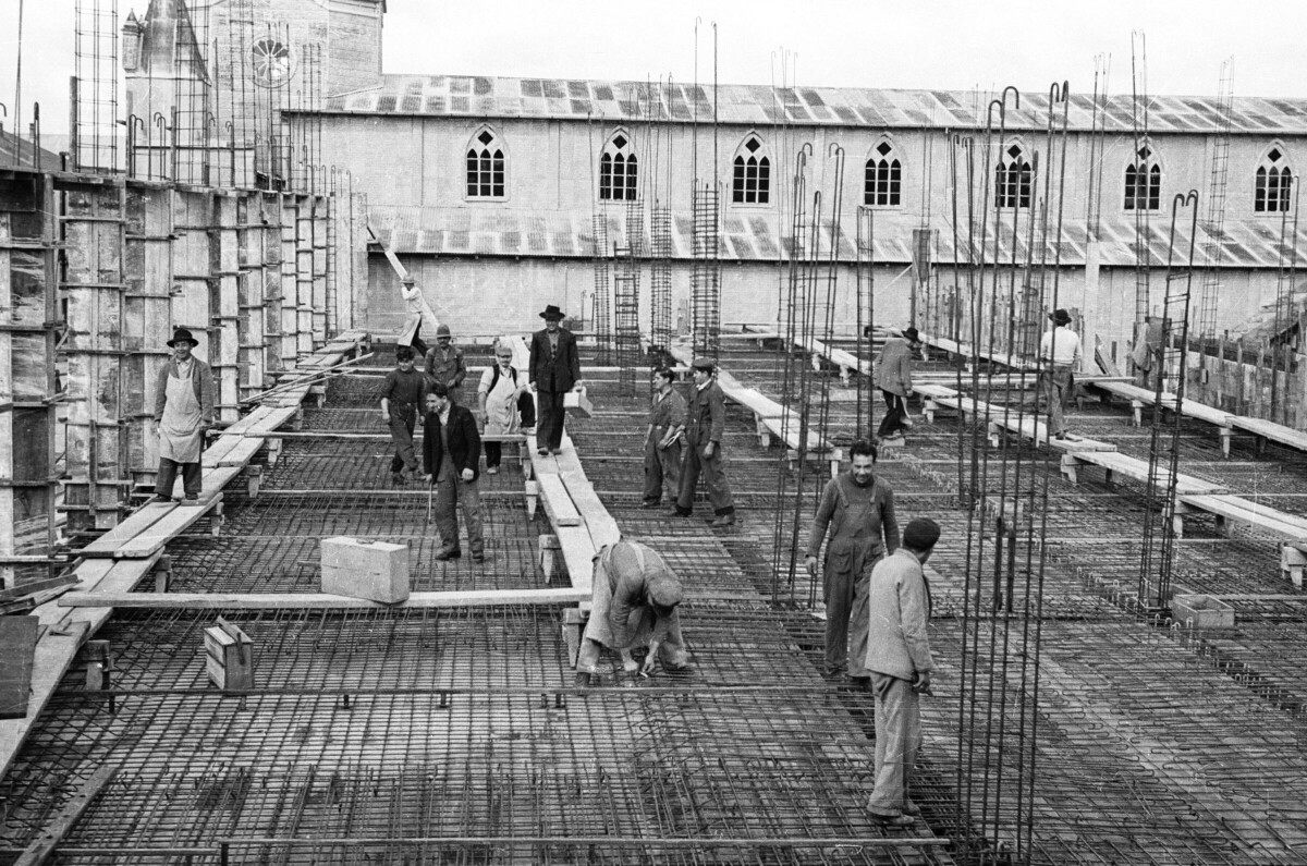 PGE de un grupo de Hombres trabajando. Dos de van caminando por encima de unos tablones de madera mientras a su alrededor los otros hombres están trabajando sobre las mallas de enfierraduras de lozas en el interior de la construcción del lado izquierdo del edificio Municipal de La Unión