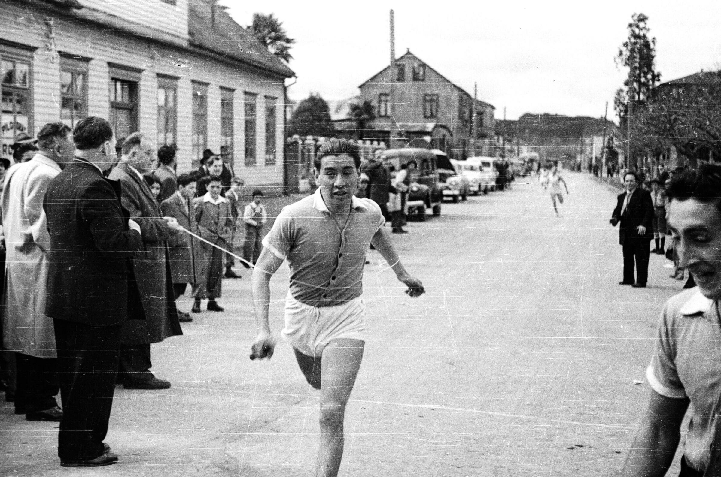 Plano entero de un hombre llegando a la meta, a la altura de su estomago tiene la cuerda de llegada la cual es sostenida por dos hombres vestidos de traje. Al final de la calle viene un corriendo un hombre también dentro de la competencia. En la meta hay un grupo de Hombres, Niños y Mujeres observándolos de pie sobre la vereda junto a la calle.