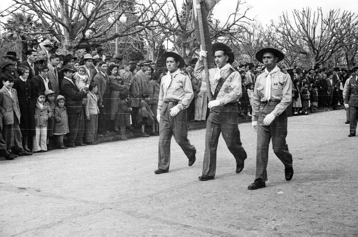 plano general de tres hombres caminando en hilera, Todos están vestidos con un uniforme compuesto por un sombrero, Camisa, pantalón, zapatos, Pañolón y guantes blancos. El hombre que camina al medio va sosteniendo el mástil de una bandera. A un costado de la calle, hay un grupo de Hombres, Mujeres, Niños y Niñas observándolos de pie sobre la vereda durante un desfile por las calles de La Unión
