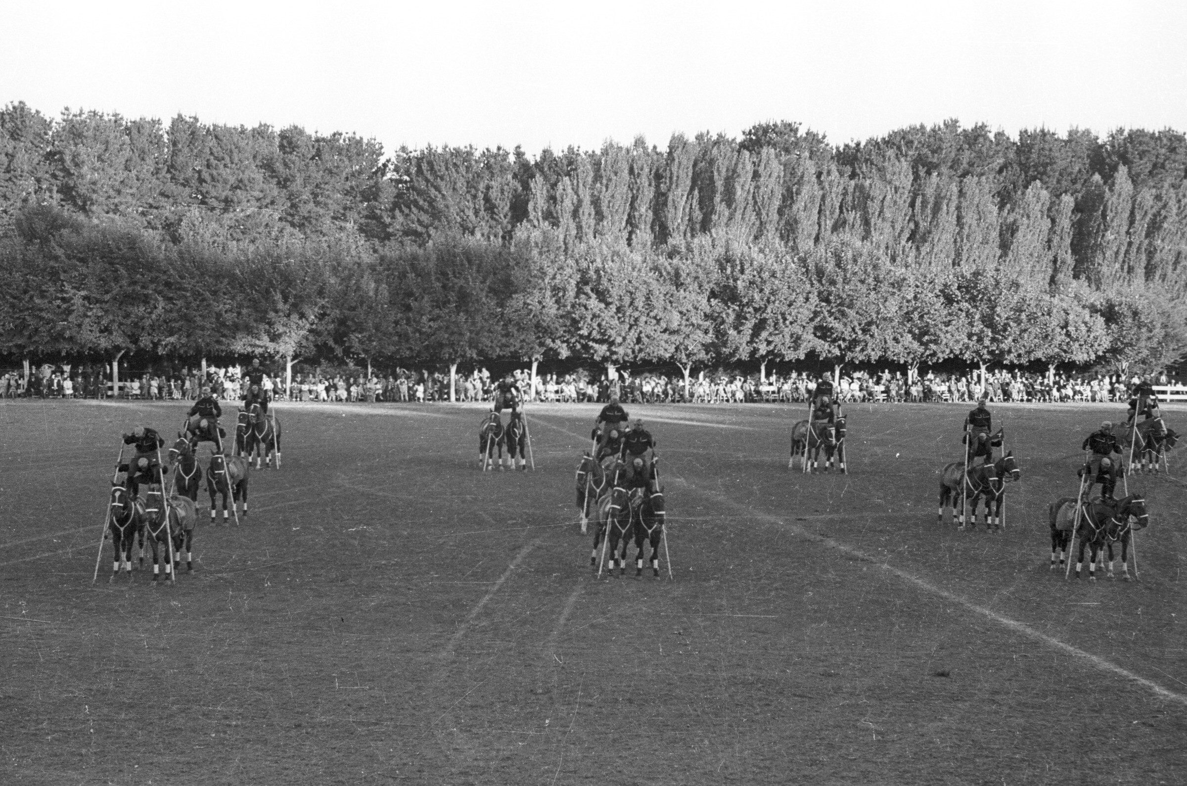 plano general de un grupo de jinetes realizando una presentación de equitación sobre los hombros de su compañero, quien a su vez se encuentra parado sobre las monturas de dos caballos. En el fondo, Niñas, Hombres y Mujer los observan sentados a un costado de la cancha ubicada en el estadio Carlos Vogel