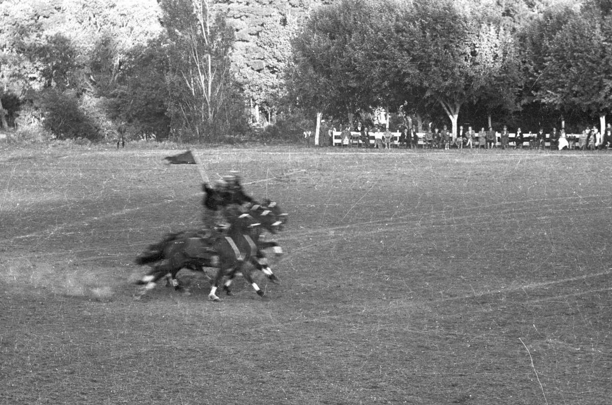 Plano entero de tres jinetes parados sobre la montura de sus caballos mientras realizan su presentación. Uno de ellos va levantando una bandera. En el fondo, Niñas, Hombres y Mujer los observan sentados a un costado de la cancha ubicada en el estadio Carlos Vogel