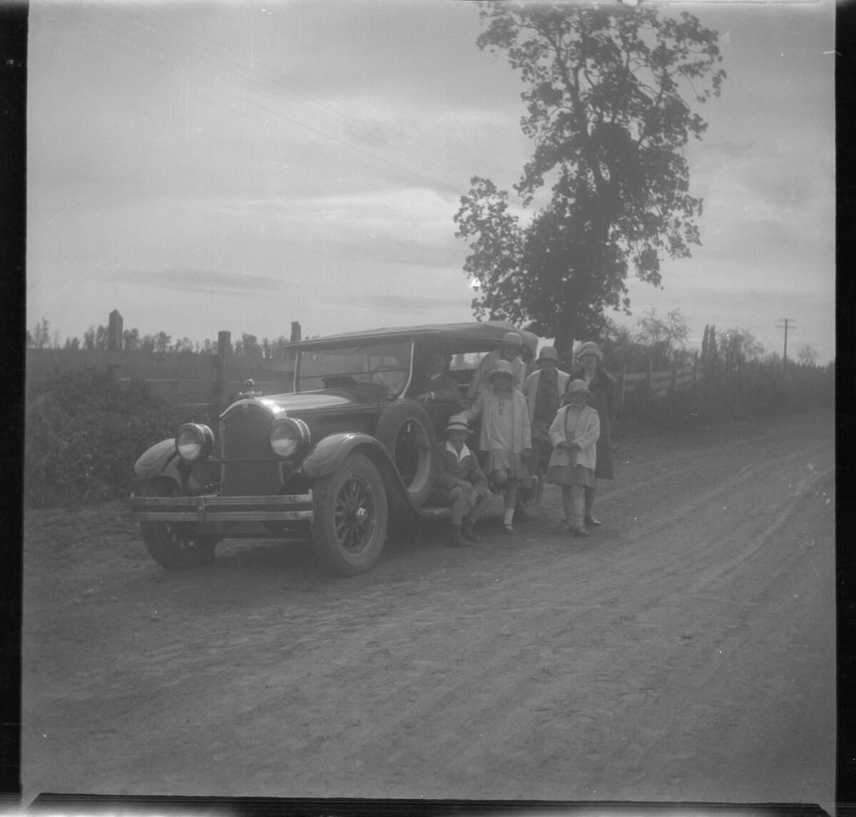 Plano general de un grupo de personas: un hombre dentro un automóvil, un niño sentado en el peldaño de la puerta del conductor, una niña que asoma su cuerpo por el automóvil, una niña apoyada en el automóvil, dos mujeres jóvenes y una niña pequeña.