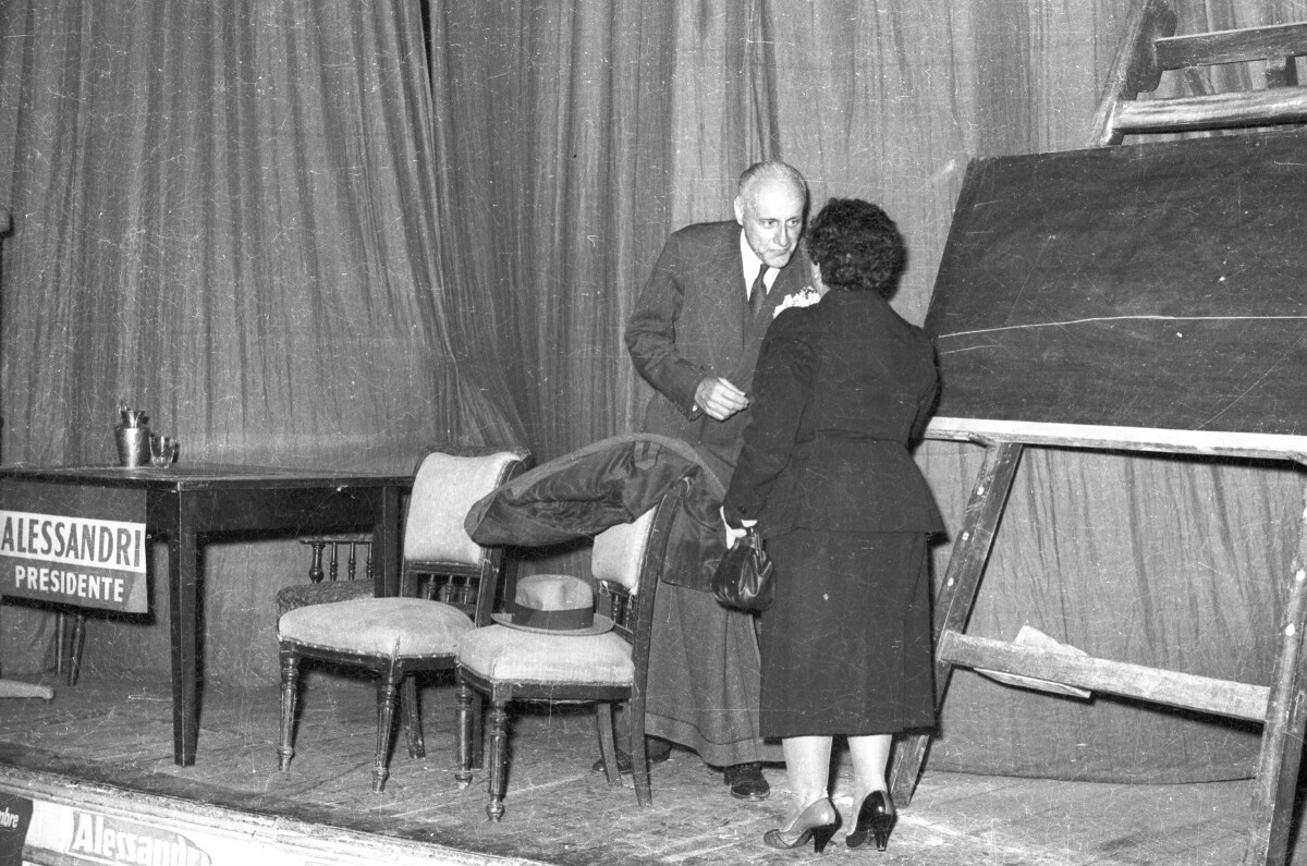 plano general de una mujer conversando con Jorge Alessandri sobre una tarima, durante su campaña presidencial realizada en la ciudad de La Unión