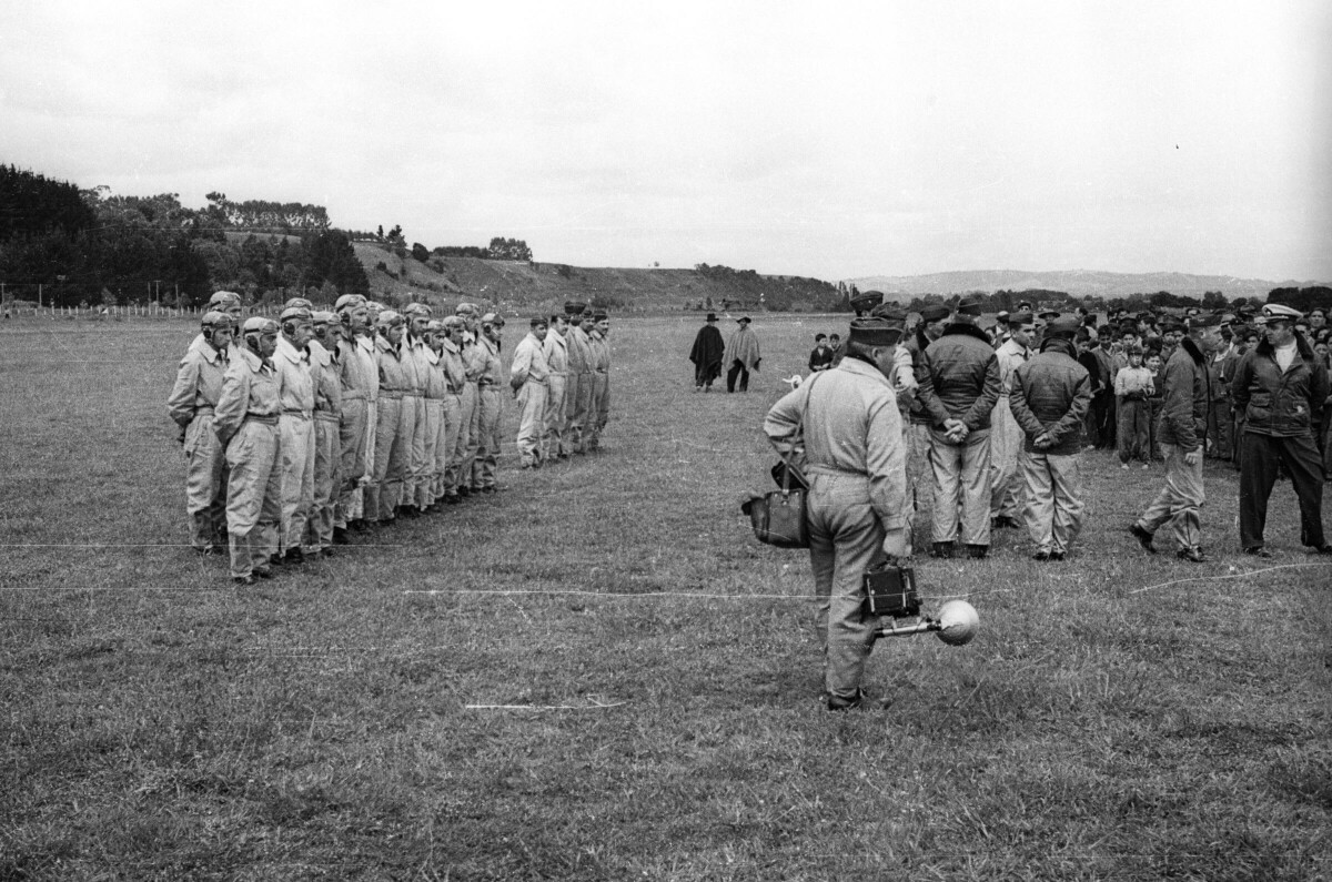 Plano entero de un grupo de aviadores formados en hileras sobre el pasto a un costado del cuadro. En el medio, uno de los aviadores va caminando de espaldas a la cámara mientras carga su equipo fotográfico. En el lugar también se encuentra un grupo numeroso de Niños, Mujer y Hombres observándolos desde un costado. Se encuentran en el interior de un aeródromo