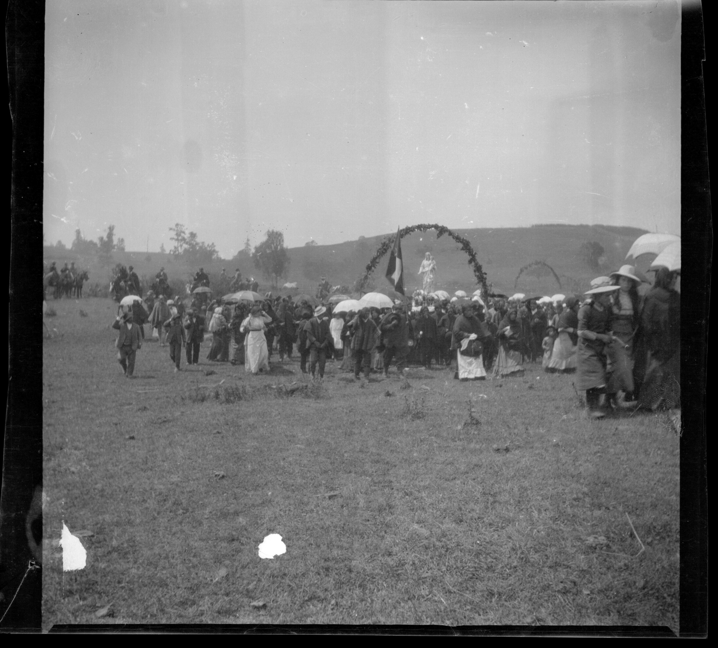 Multitud de personas en ceremonia religiosa, procesión. Algunas personas sostienen paraguas.