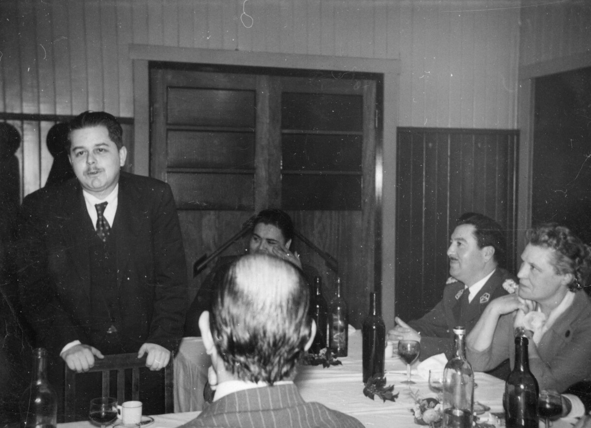 plano general de un hombre vestido de Traje, parado detrás de una silla mientras se dirige al resto de los invitados que están sentados alrededor de las mesas. En un costado se encuentra Olga Boettcher sentada mirándolo mientras afirma su barbilla sobre ambas manos. Se encuentran en el interior de una infraestructura no identificada durante una celebración