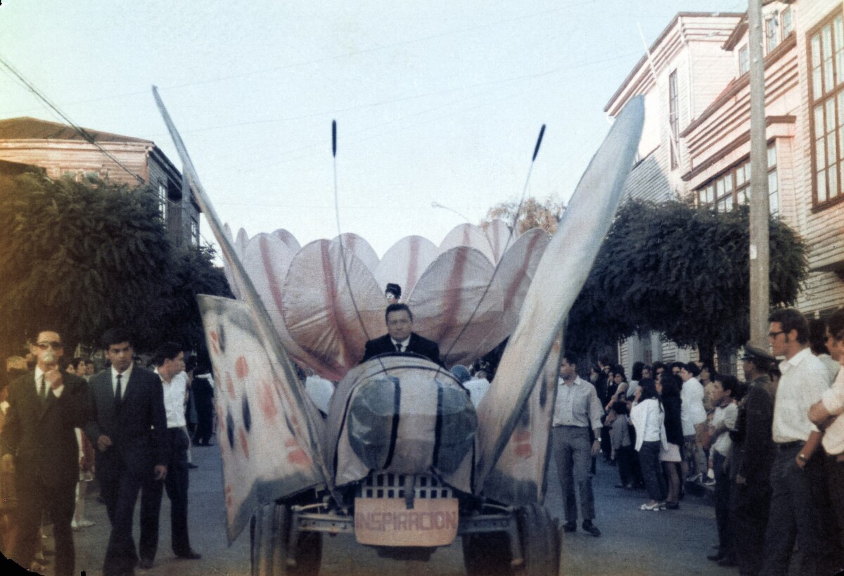 Plano entero de un carro alegórico de mariposa y una flor con un cartel que dice -Inspiración- paseando por una calle de La Unión