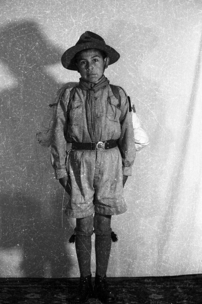Retrato de estudio de Plano entero de un niño posando de frente. Pertenece a una brigada no identificada y lleva puesto su uniforme, un sombrero y una mochila
