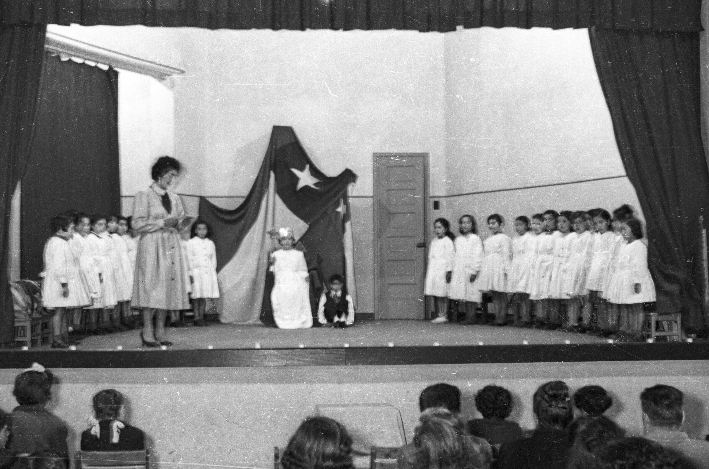 plano general de una mujer con vestido, leyendo un papel, parada sobre un escenario. En el fondo un grupo de Niñas se encuentran paradas junto a sus sillas mientras que más en el fondo, una niña y un niño se encuentran sentados bajo una bandera chilena la cual se encuentra colgada en la pared. Se encuentran realizando una presentación artística en el gimnasio de la escuela de Niñas Nº2