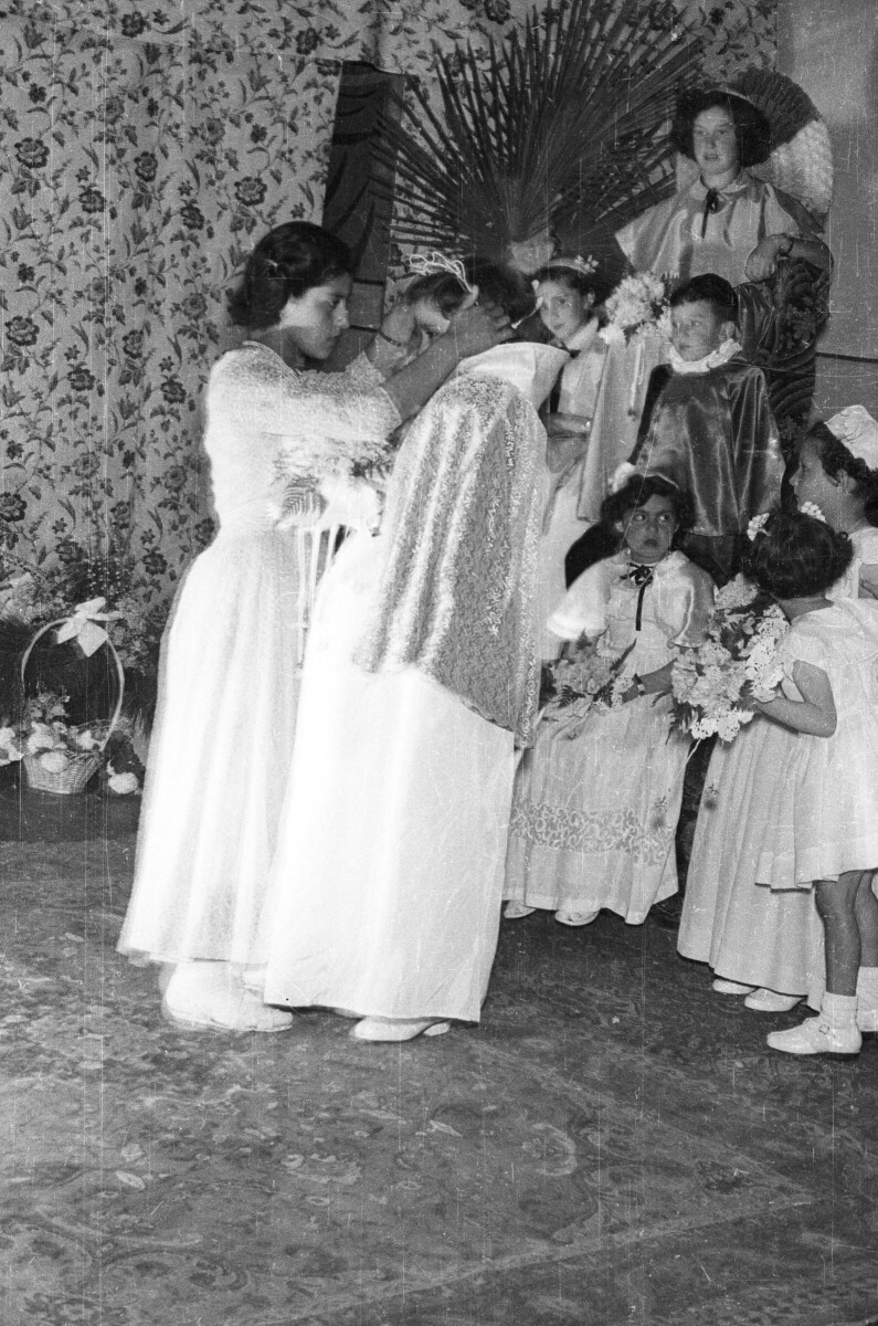 Plano entero de un niña colorando una corona sobre la cabeza de otra Niña. En el fondo hay un grupo de Niñas sentadas con un ramo de flores y sobre un asiento de gran altura hay una Niña sentada observando con un Niño junto a ella. Están sobre el escenario durante la coronación de la reina durante una jornada de presentaciones artísticas en el interior de una escuela.