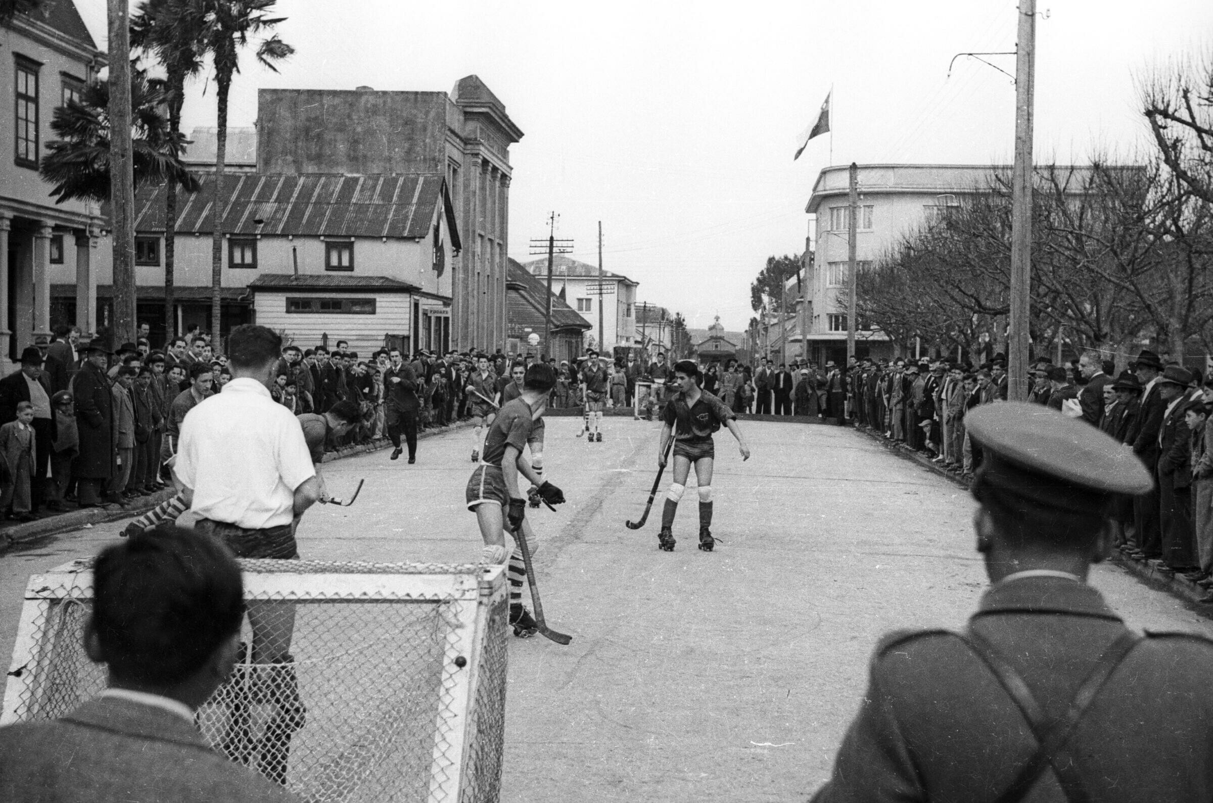 plano general de dos equipos de hockey masculinos no identificados, jugando un partido sobre una calle pavimentada, la cual se encuentra cortada por un grupo numeroso de personas que observan paradas alrededor de los jugadores, por sobre la vereda.