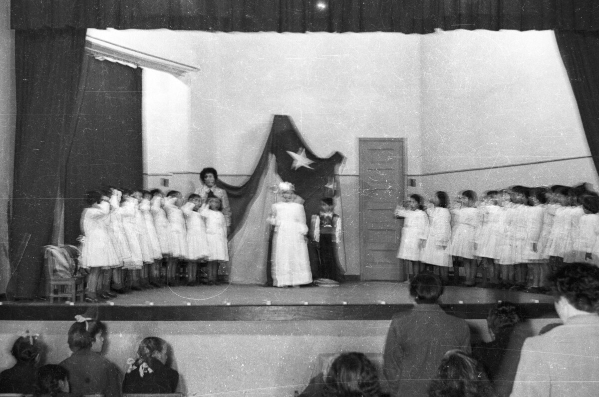 plano general de dos grupos de Niñas, cantando con una mano a la altura de su cabeza. Junto a ellas una Niña vestida de reina, un niño y una mujer los acompañan sobre el escenario. En el fondo, una bandera chilena se encuentra colgada en la pared. Están realizando una presentación artística en el gimnasio de la escuela de Niñas Nº2