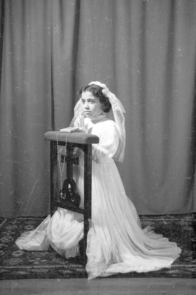 Retrato de estudio de Plano entero de niña con Vestido de primera comunión arrodillada en reclinatorio con rosario y manos juntas.