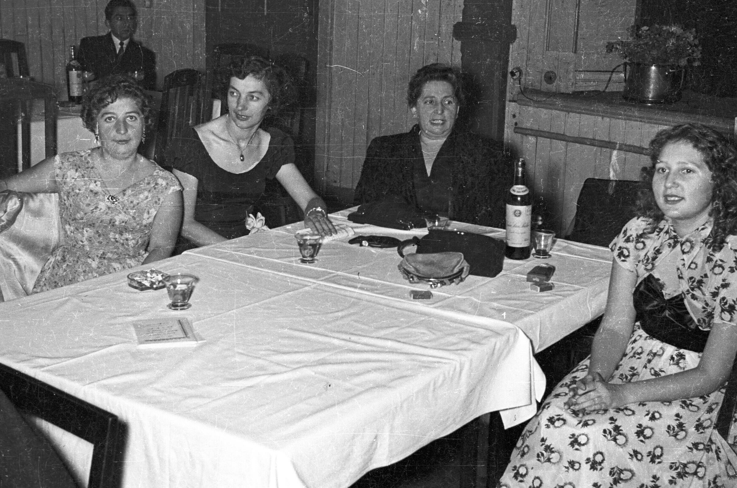 Plano entero de cuatro mujeres con Vestidos sentadas alrededor de una mesa, sobre la cual tienen sus carteras y una botella de licor. Una de ellas se encuentra mirando hacia la cámara. Se encuentran en el salón del Club Alemán de La Unión, durante una fiesta