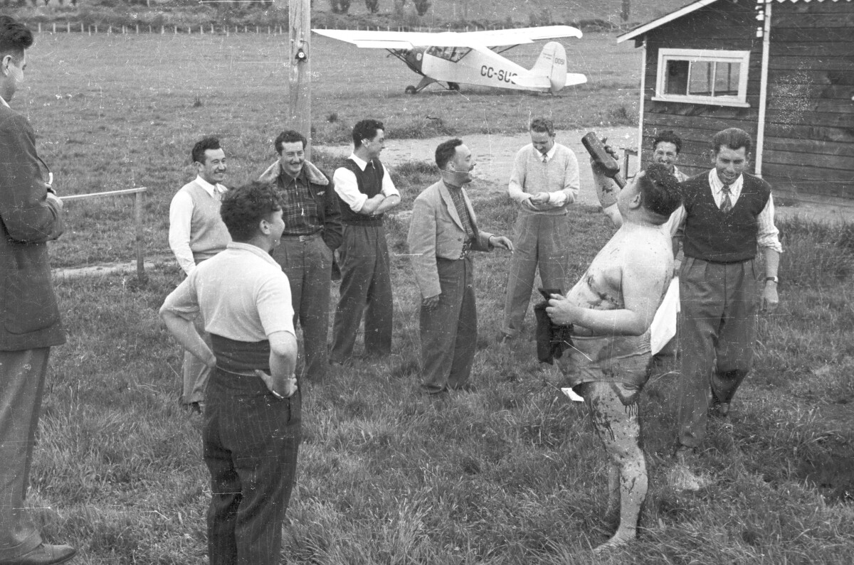 plano general de un hombre semidesnudo, bebiendo de una botella empinada. A su alrededor un grupo de Hombres lo acompañan mientras ríen y conversan. Están ubicados junto a una casa. En el fondo, hay una avioneta estacionada. Están celebrando el rito de iniciación del nuevo integrante en el interior del club aéreo de La Unión