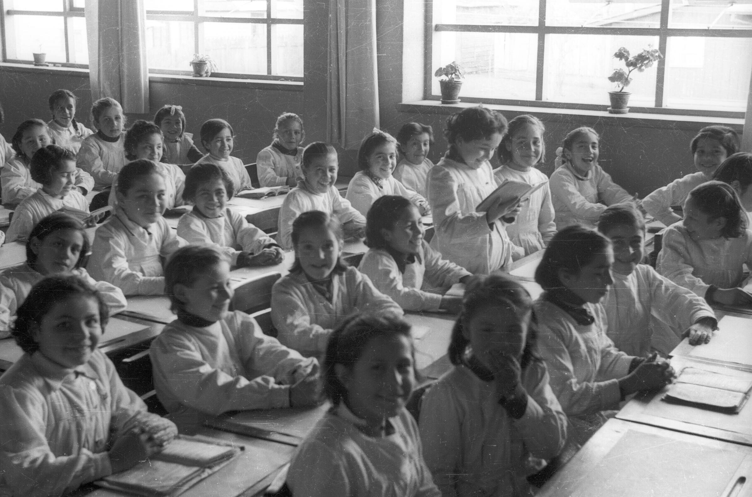plano general de un grupo numeroso de niñas sonriendo mientras está sentadas frente a sus pupitres sobre el cual tienen un libro abierto. Se encuentran en una de las salas de la escuela superior de niñas Nº2