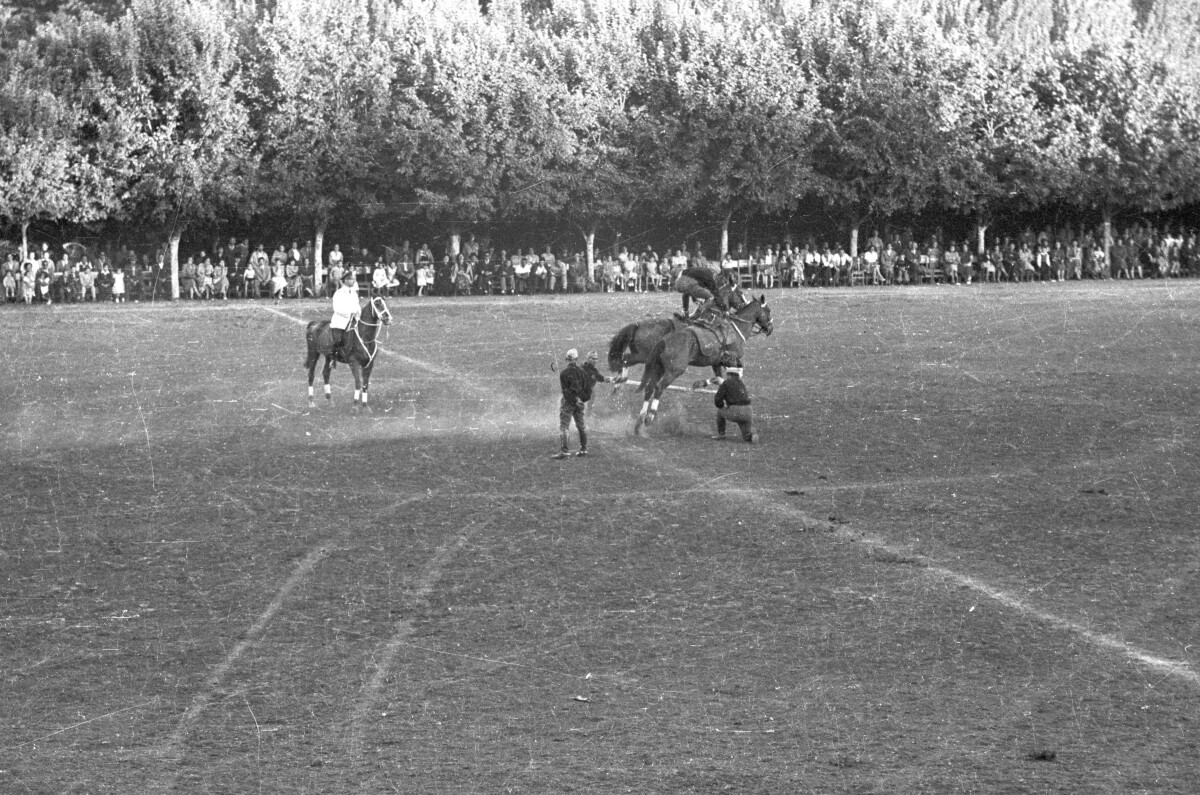 Plano entero de un hombre parado en dos caballos, saltando sobre una valla sostenida por otros dos hombres. En un costado, un carabinero montado a caballo los observa mientras realizan su presentación. En el fondo, Niñas, Hombres y Mujer los observan sentados a un costado de la cancha ubicada en el estadio Carlos Vogel
