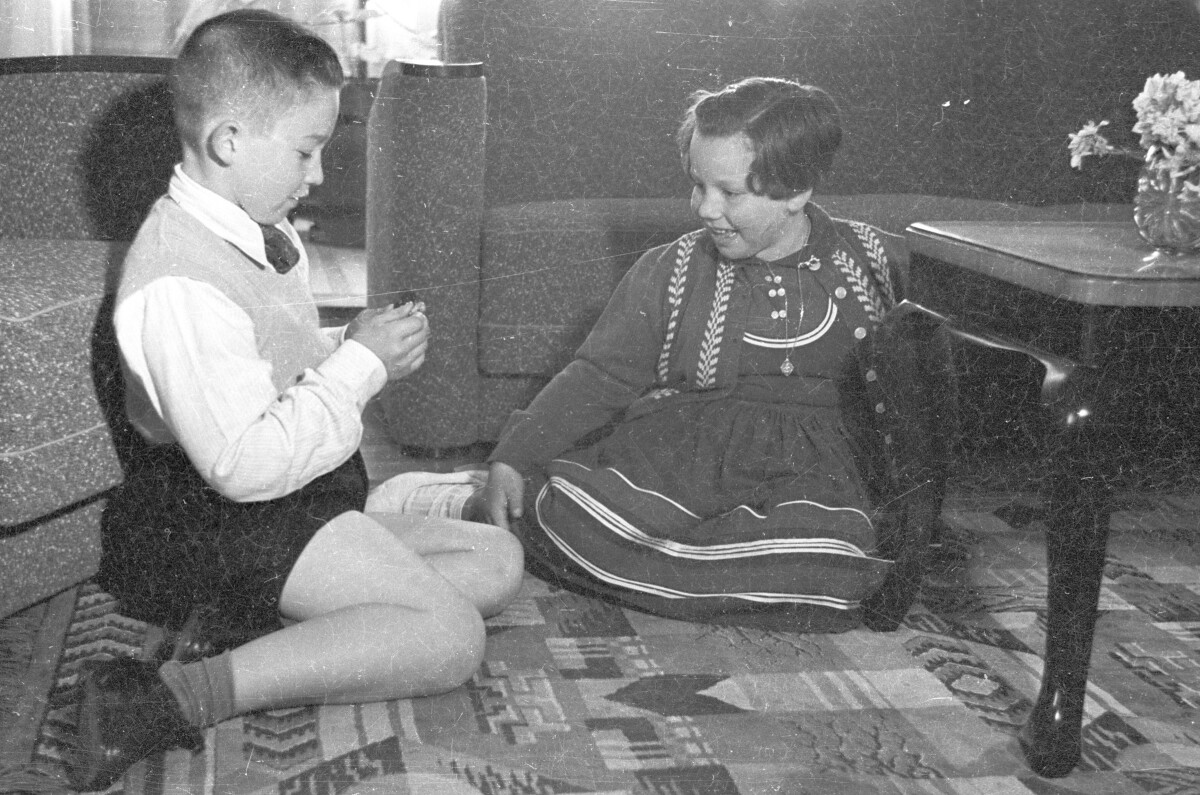 Plano entero de un niño observando un objeto identificado en compañía de una Niña. Ambos se encuentran sentados sobre una alfombra en el interior de la sala de una casa