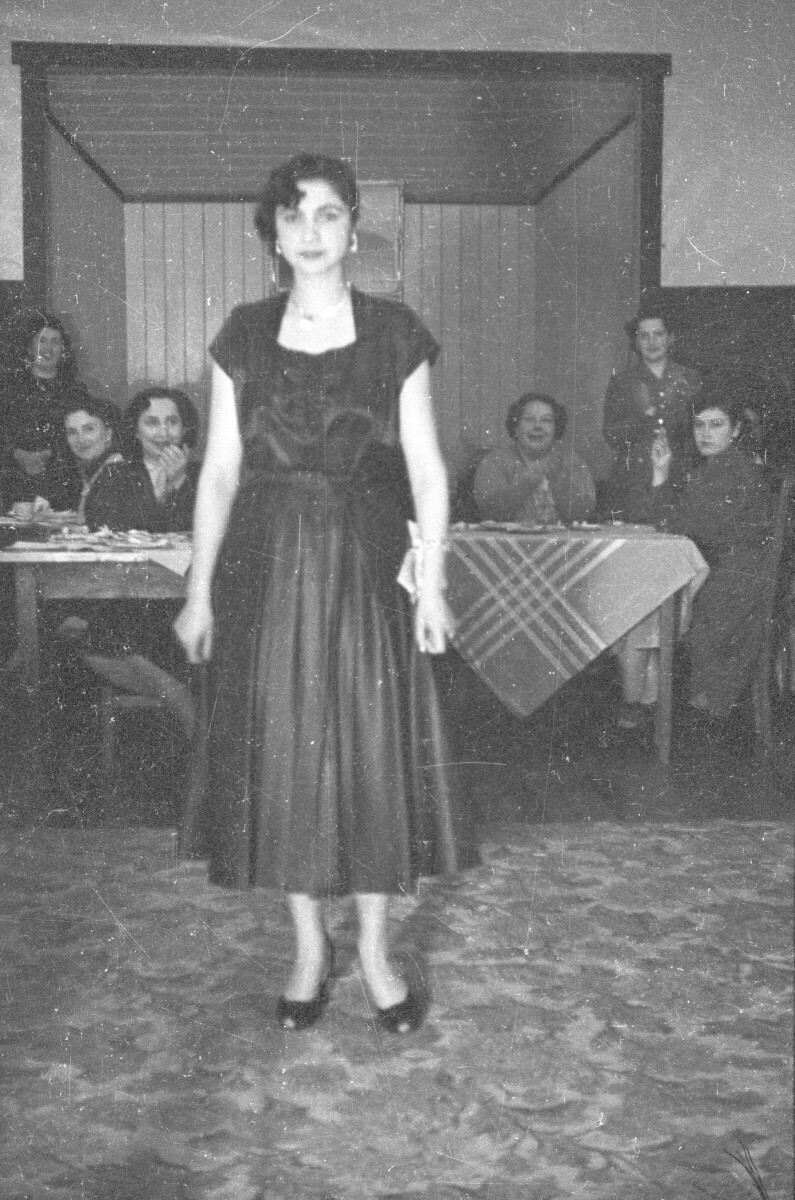Plano entero de una mujer modelando un vestido, en el fondo hay un grupo de mujeres que la aplauden mientras la observan sentadas alrededor de unas mesas en el interior de un local