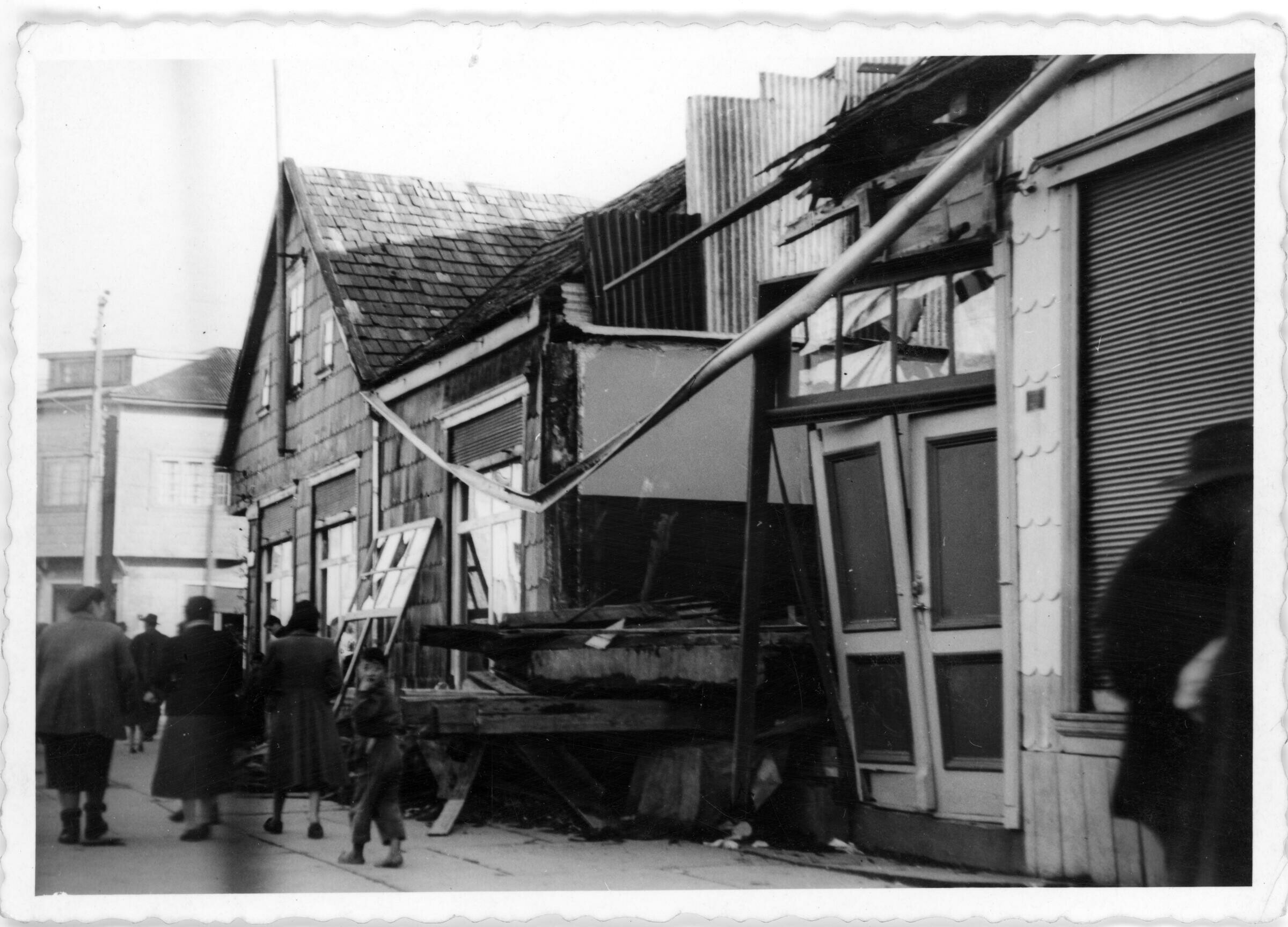Plano general de un grupo de personas caminando en el frontis de una casa de madera derrumbada por el terremoto de 1960 en el centro de Puerto Varas.
