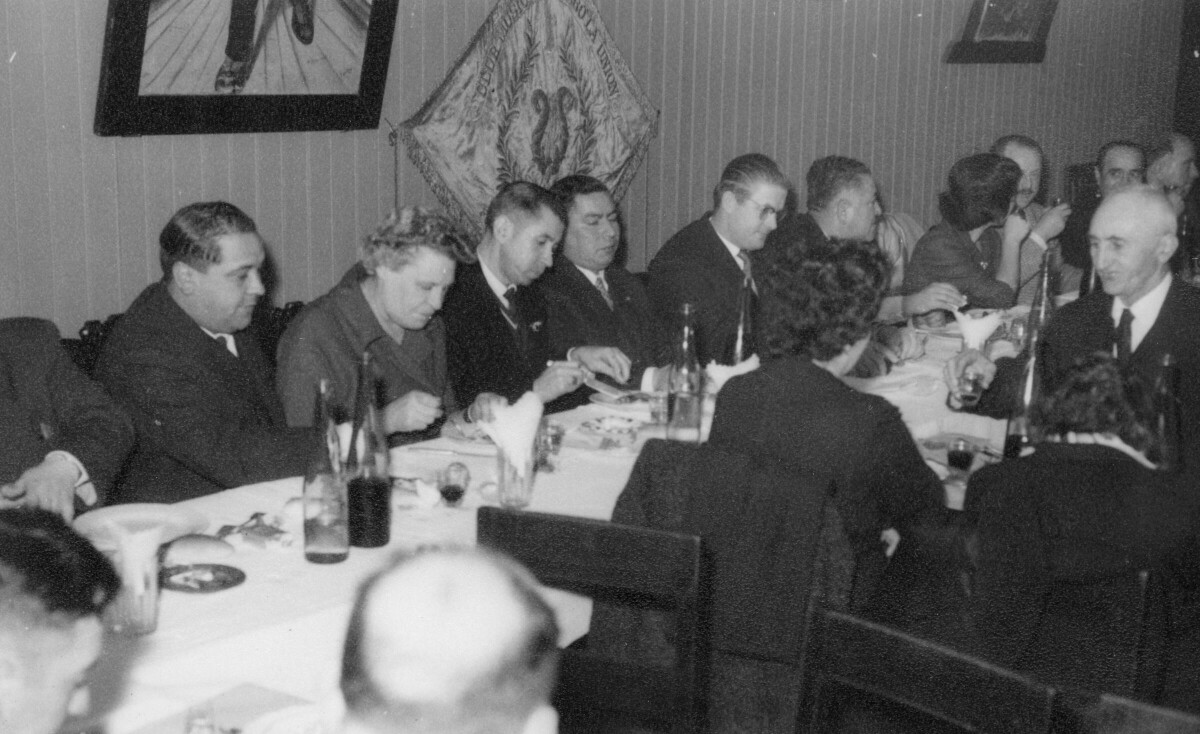 Plano entero de un grupo de Hombres y Mujeres, sentados alrededor de largas mesas durante un banquete. La primera mujer de izquierda a derecha es Olga Boettcher, primera gobernadora de la ciudad de La Unión. Se encuentran en el interior de una infraestructura no identificada durante una celebración