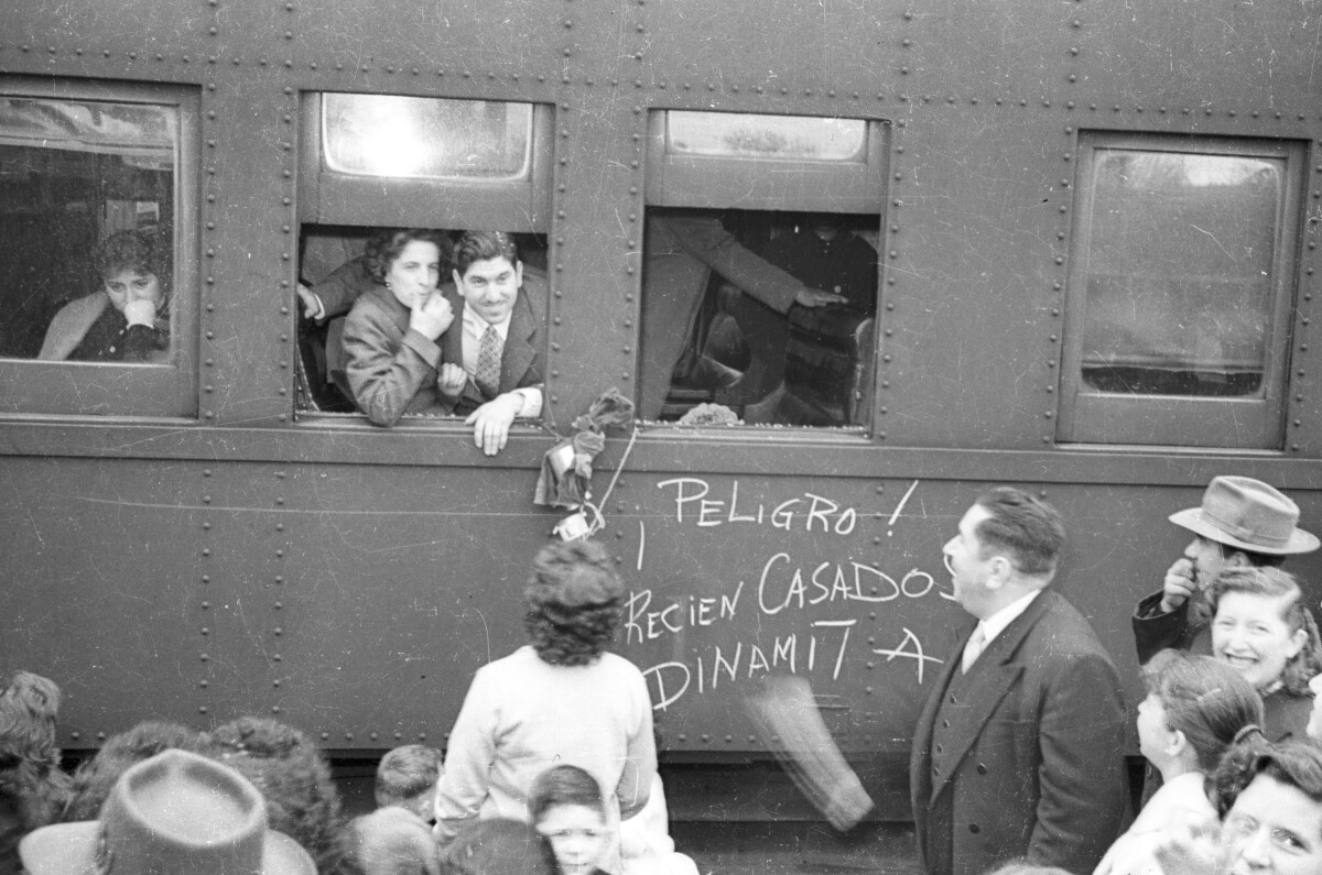 Plano entero de un matrimonio conversando con un grupo de Mujeres y Hombres desde la ventanilla del tren, en el cual está escrita la frase -¡Peligro! recién casados Dinamita!-. Están a prontas de partir a su luna de miel. Se encuentran en la estación de trenes de La Unión
