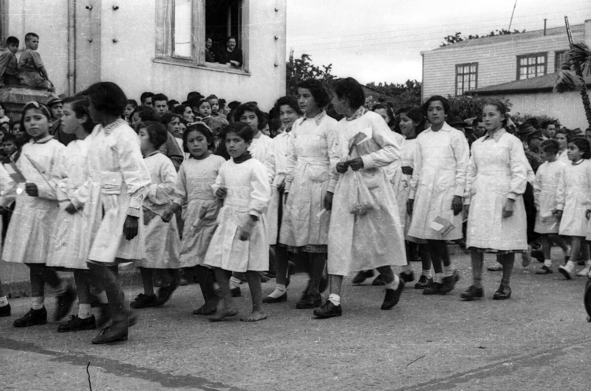 Plano entero de un grupo de niñas con delantal, desfilando formadas en hileras. En el fondo, un grupo de personas se encuentran observándolas en el frontis de la municipalidad