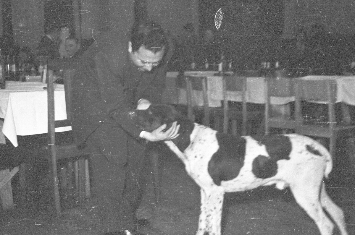 plano general de un hombre acariciando a un perro blanco con manchas negras, durante una celebración dentro de un local.