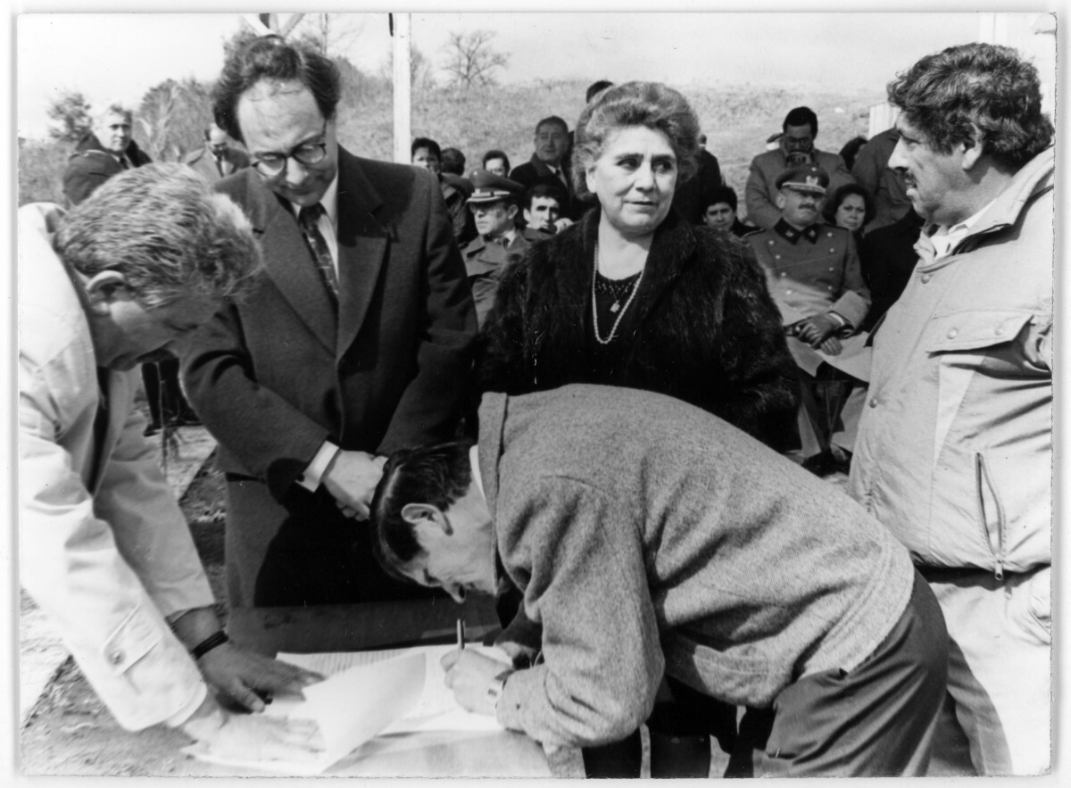 Plano entero de un hombre agachado firmando un libro sobre una mesa mientras dos hombres a su lado lo observan. Junto a ellos se encuentra Elfrida Ojeda mirando al otro hombre que está parado a su lado. En el fondo, hay un grupo de Hombres, Mujeres y dos oficiales de carabineros sentados sobre una tarima. Se encuentran en el interior de un recinto no identificado durante la entrega de unas viviendas sociales