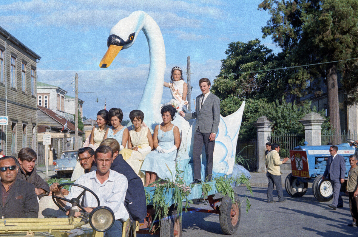 plano general de la reina de la primavera, sonriendo a la cámara junto a un grupo de Hombres y Mujeres desde un carro alegórico caracterizado como un cisne, el cual se desplaza por las calles de la Unión