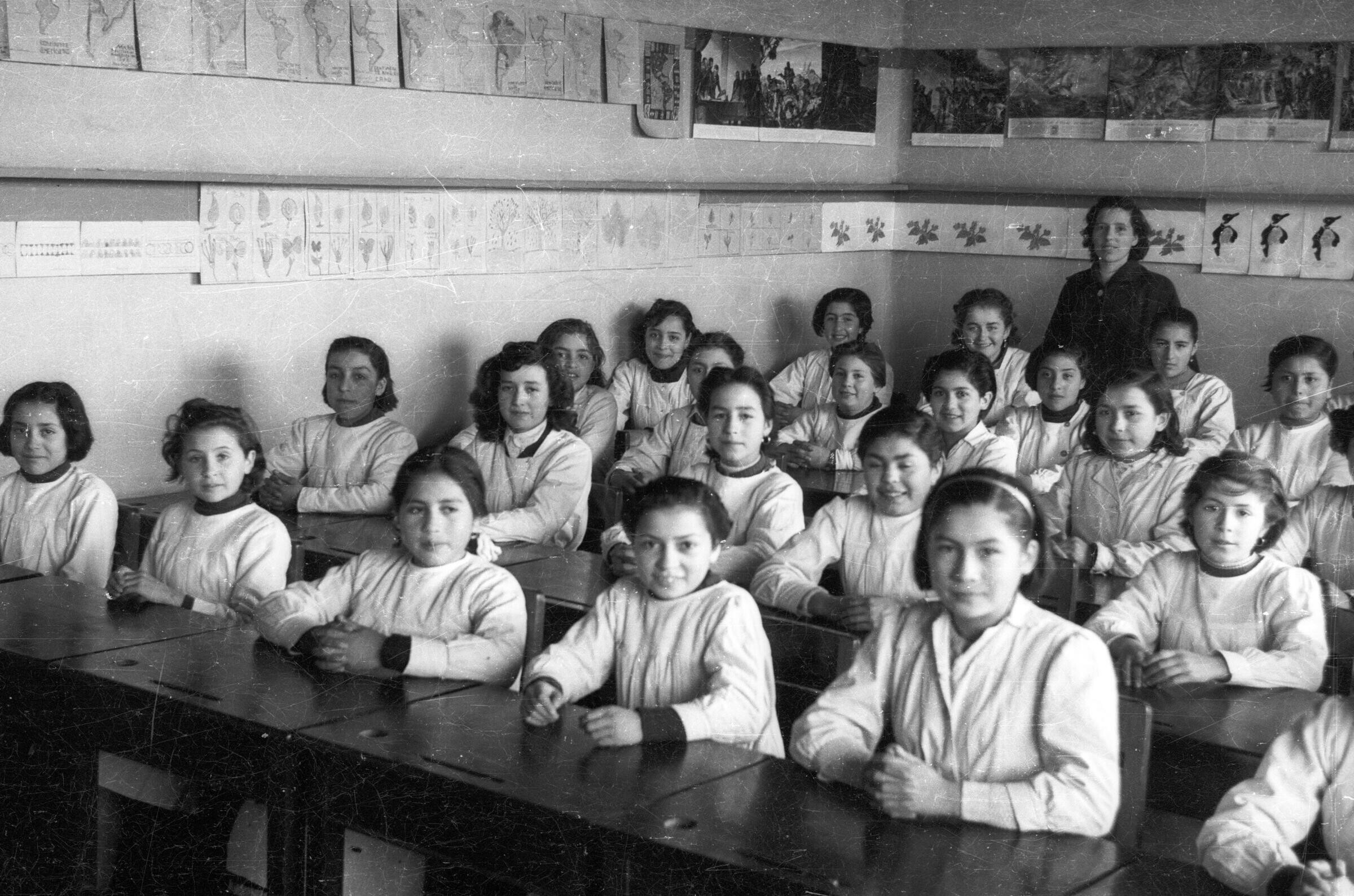 Retrato grupal de plano entero de niñas posando sentadas frente a sus pupitres y una profesora en el fondo de la sala de la escuela superior de niñas Nº2