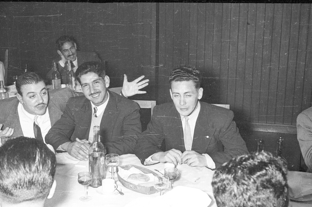 Plano medio de un hombre posando mientras sonríe, junto a el hay dos hombres conversando sentados alrededor de una mesa durante una celebración en el interior de un local