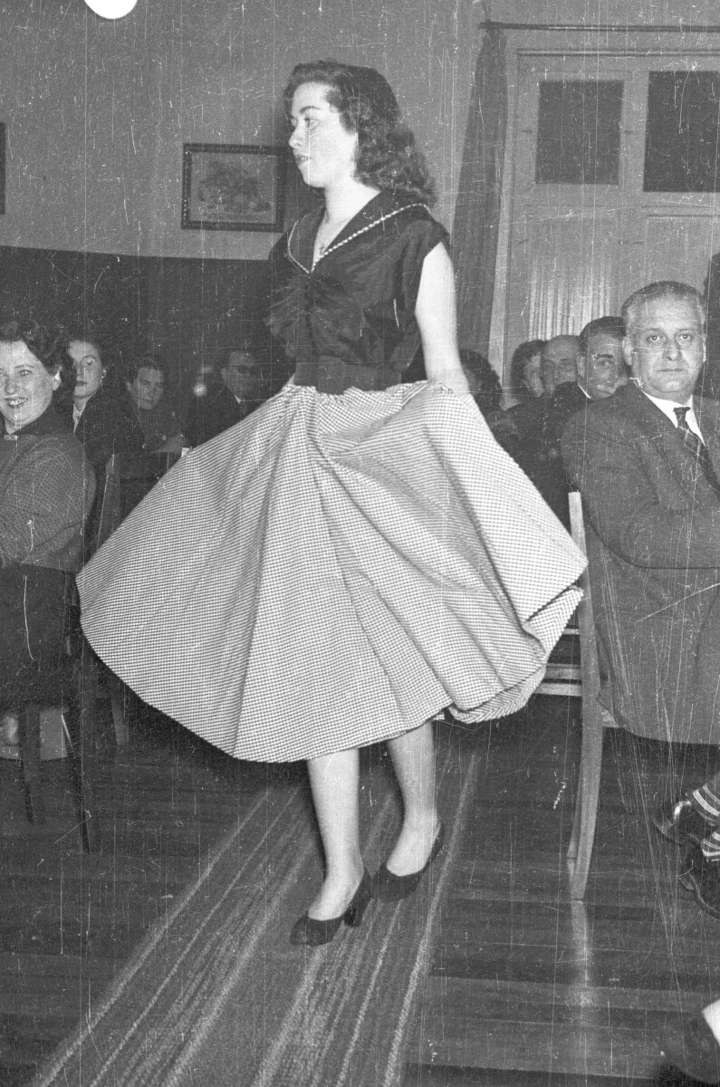 Plano entero de una mujer modelando un Vestido mientras levanta con ambas manos su falda, va caminando sobre una alfombra y se encuentra mirando hacia un costado. A su alrededor hay un grupo de Mujeres y Hombres que la están sentados junto a unas mesas, que la observan. Están en el interior de un local durante una exhibición de Vestidos
