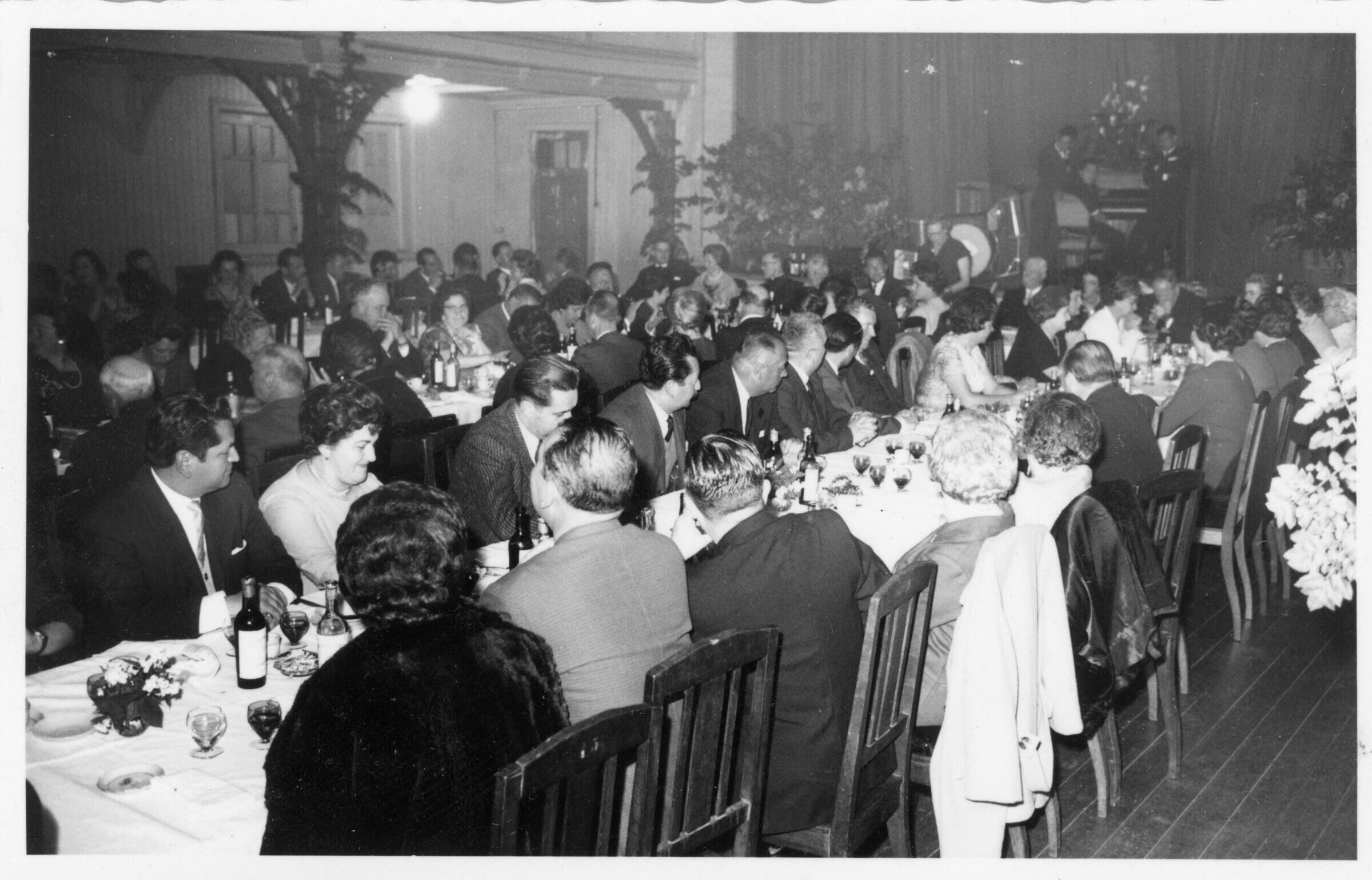 plano general de un grupo de Hombres y Mujeres sentados alrededor de largas mesas mientras comen y conversan entre sí. En el fondo, hay un grupo musical preparándose sobre el escenario. Están en el interior de un local no identificado durante un banquete realizado en honor a Olga Boettcher tras haber finalizado su gobernación