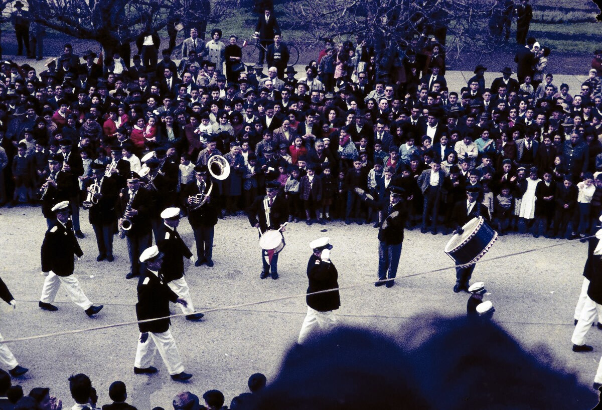 Plano entero de ejército de la Fuerza Naval desfilando junto a una banda tocando instrumentos. A los costados gran cantidad de personas observando en calle aledaña a plaza de la Concordia de La Unión