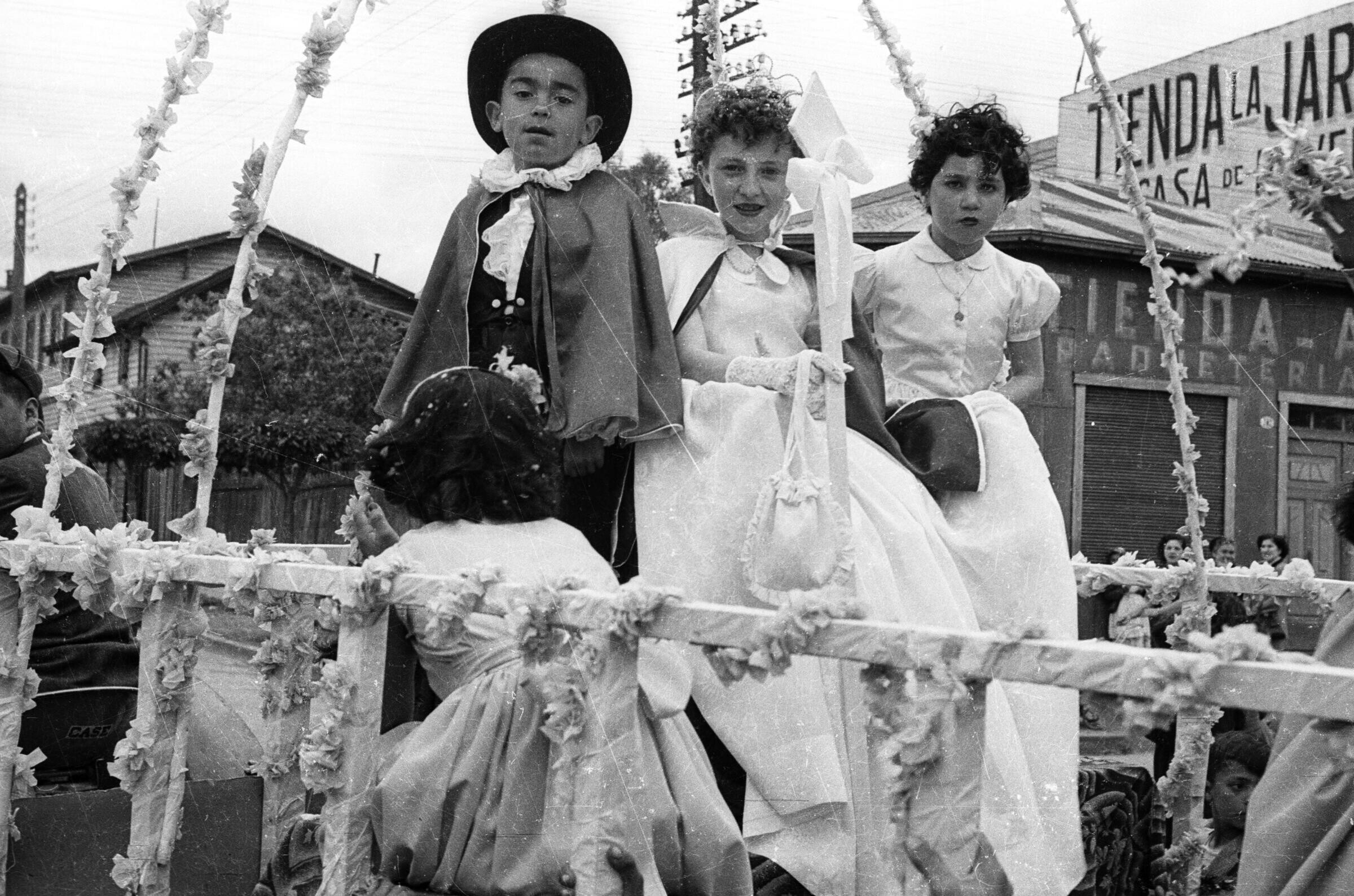 -Plano entero de una Niña y la pareja de reyes de la escuela de Niñas Nº2 mirando hacia la cámara. Se encuentra en el interior de un carro alegórico, el cual avanza por afuera de una tienda llamada --La Jardinera---
