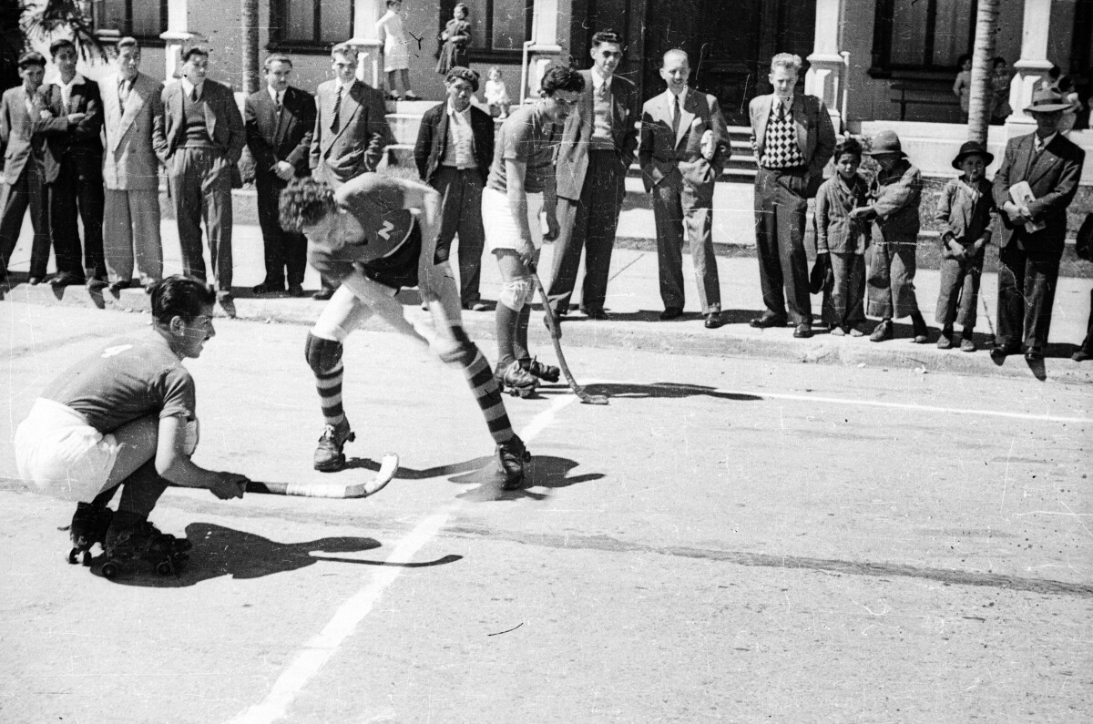 Plano entero de tres hombres jugando hockey en patines, en el fondo un grupo de hombres vestidos de traje y Niños los observan jugar a la mitad de la calle en las afueras de un edificio.