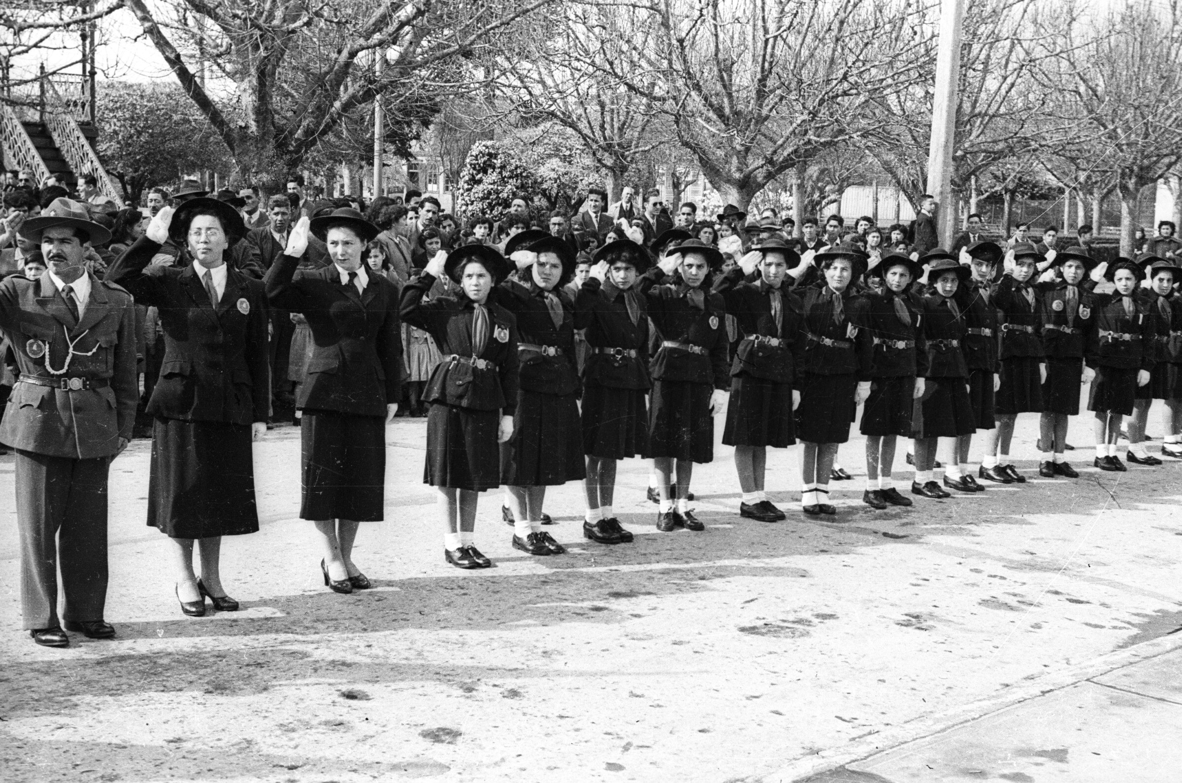 Plano entero de un grupo de mujeres, jóvenes y adultas, formadas en dos hileras mientras realizan un saludo mirando hacia su derecha. En un costado las acompaña un hombre. Todos son pertenecientes a la brigada de scout y llevan puesto su uniforme formal. En el fondo, sobre la vereda, hay un grupo numeroso de Niños, Niñas, Mujer y Hombres que observan el desfile que se está llevando a cabo en un costado de la plaza de La Concordia
