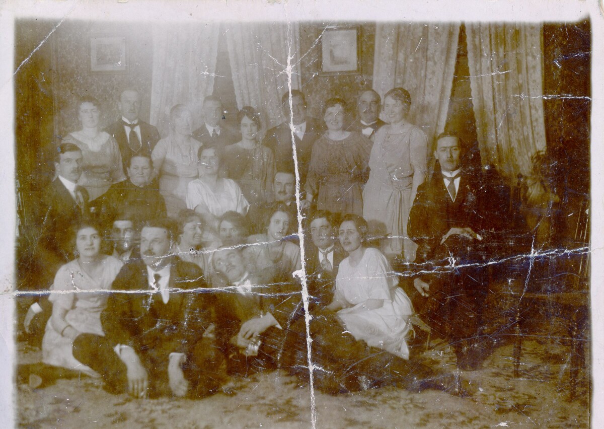 Retrato grupal de plano entero de Mujeres y Hombres posando. Algunos se encuentran sentados sobre la alfombra mientras que el resto están de pie o sentados en las sillas. Se encuentran en el interior de una casa