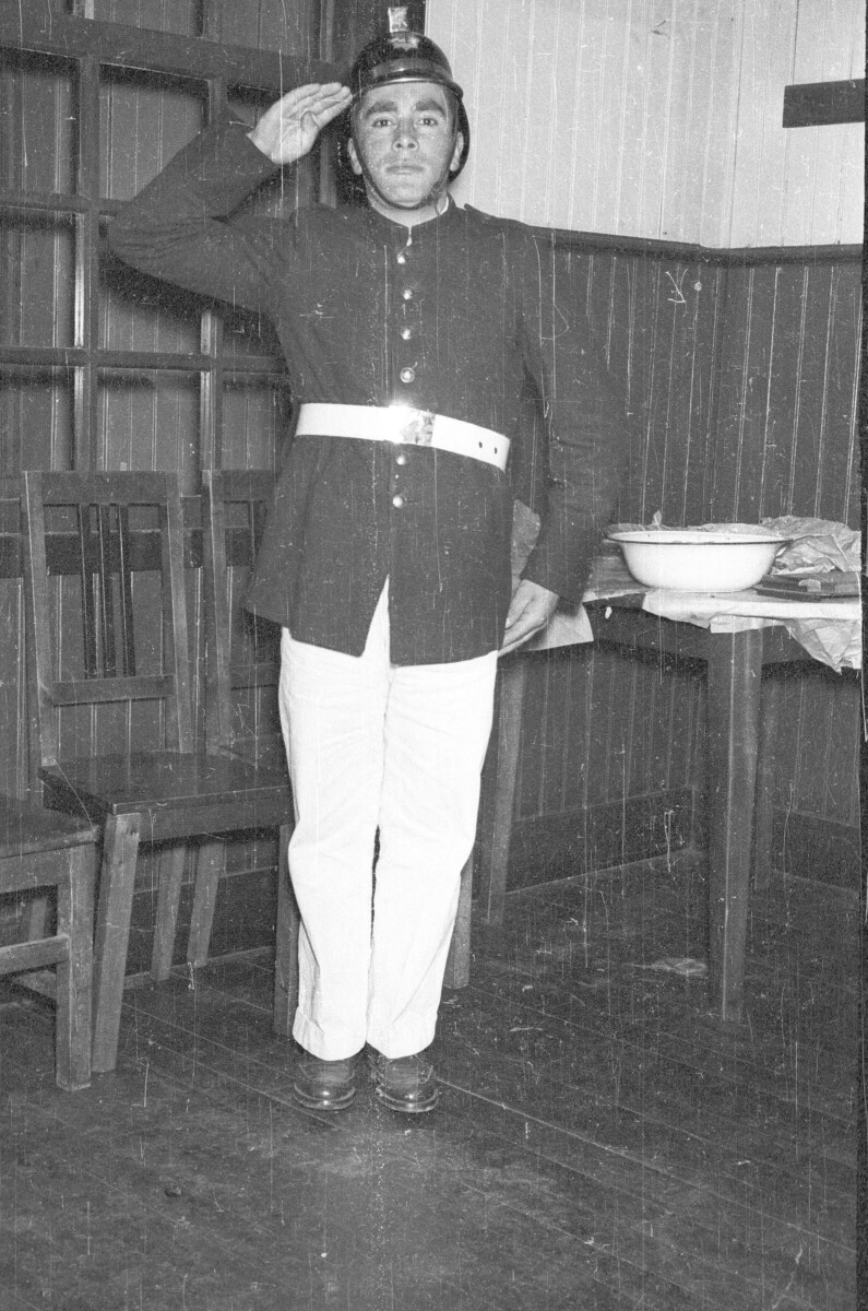 Retrato de plano entero de un hombre posando de pie mientras realiza un saludo a la altura de la cabeza. Está usando su uniforme de bombero junto a una mesa con una fuente encima. Está en el interior de una infraestructura.
