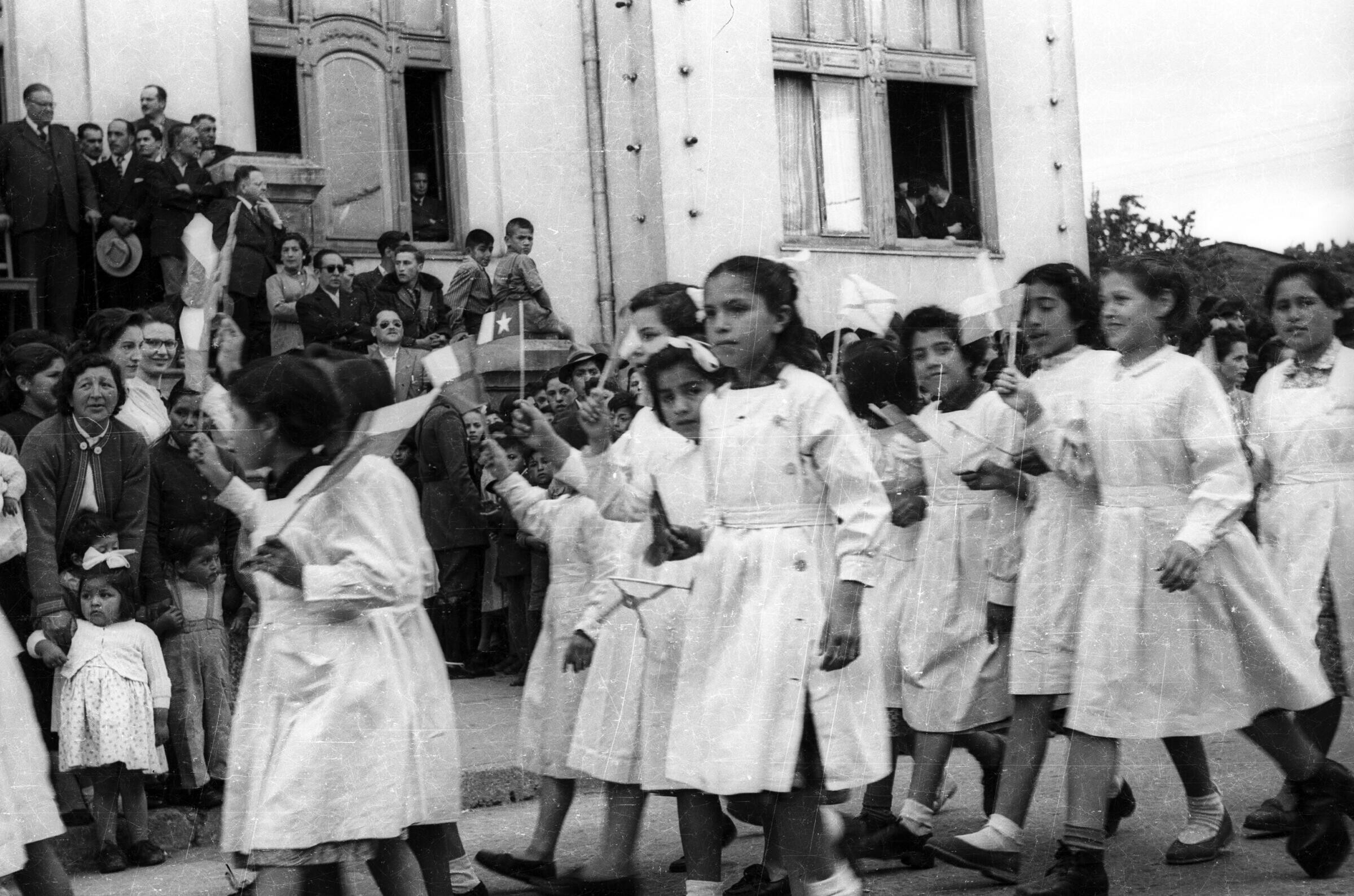 plano general de un grupo de niñas con delantal, desfilando formadas en hileras, con una bandera en las manos. En el fondo, un grupo de personas y Carlos Vogel se encuentran observándolas en el frontis de la municipalidad